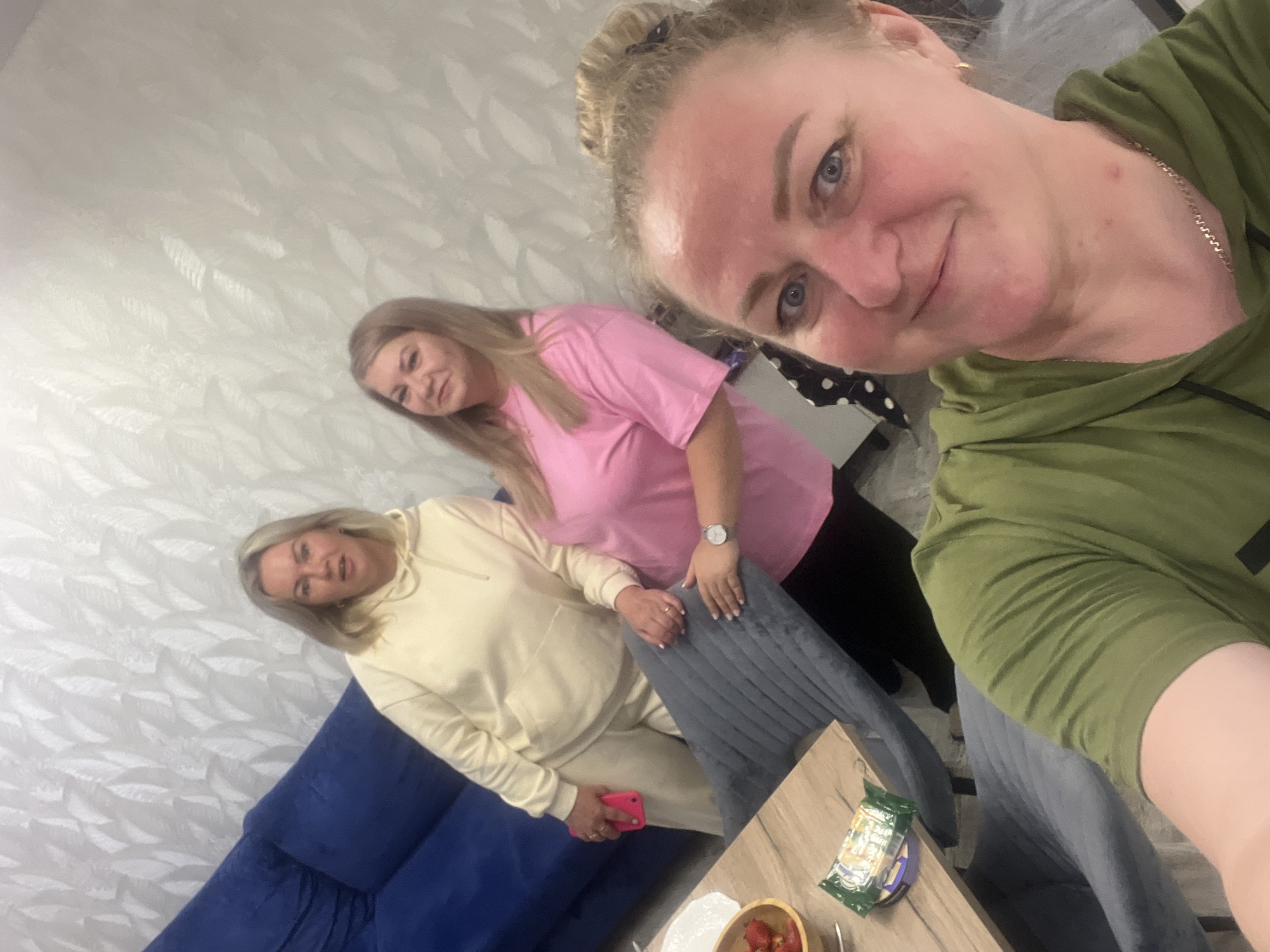 Three adult females are captured in a selfie taken indoors in Старые Кены, Russia. The individual in the foreground, with blonde hair in a bun and wearing a green hooded sweatshirt and a gold chain, is taking the photo, looking at the camera with a slight smile. Behind her, a female with light brown hair and a pink t-shirt smiles towards the camera, her left hand resting on a grey upholstered dining chair. She wears a silver watch on her left wrist. Adjacent to her, a third female with blonde hair, wearing a light yellow hooded tracksuit and holding a bright pink smartphone in her left hand, also looks towards the camera with a neutral expression. The background wall features a textured, light-colored wallpaper with a subtle leaf-like pattern. A blue upholstered sofa is partially visible on the far left. A wooden table in the mid-ground holds a bowl of strawberries, a white plate, a green packaged item, and a small purple container. The setting appears to be a modern residential interior.