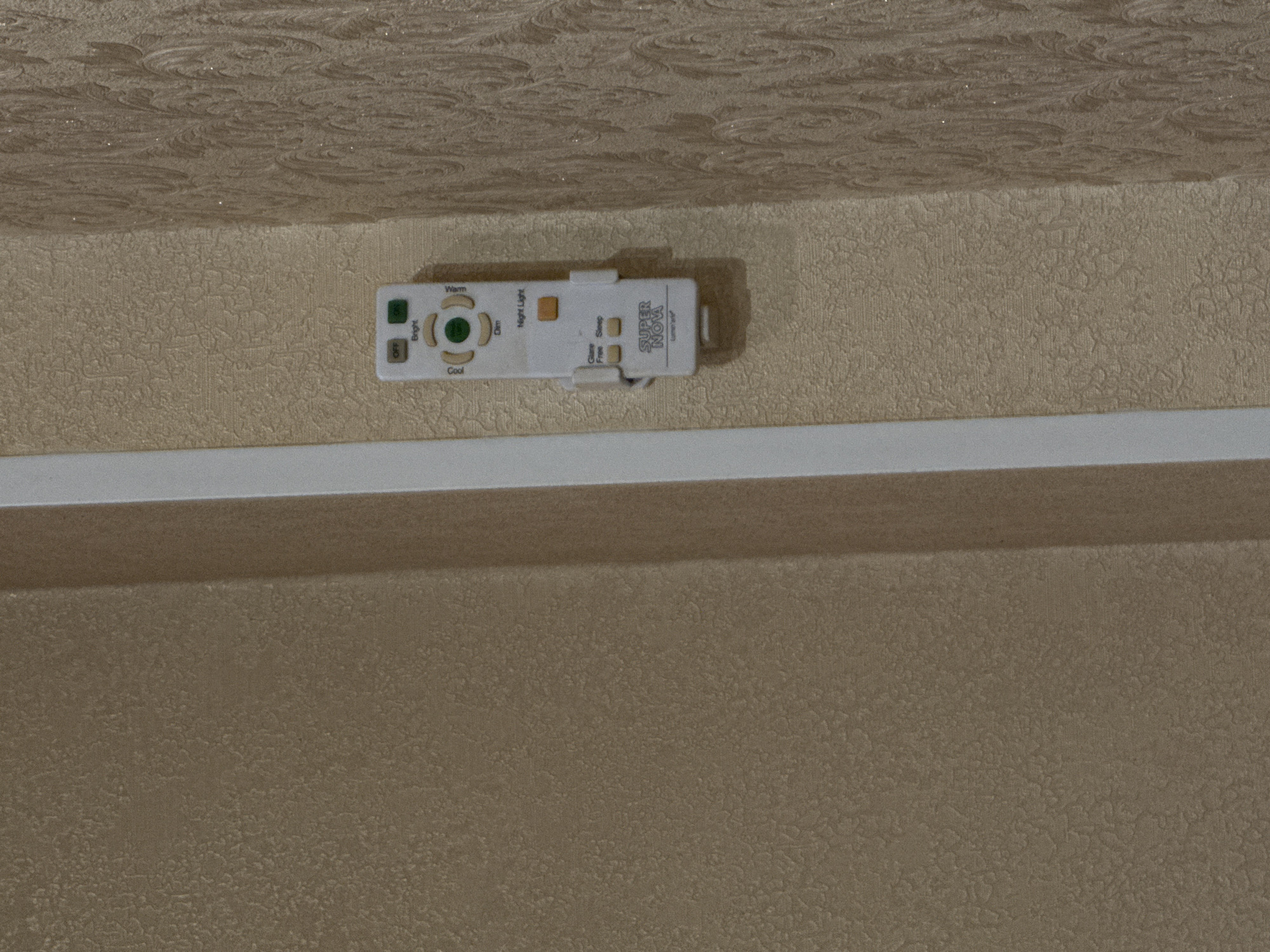 A white remote control is mounted on a textured beige wall, vertically aligned. The remote has several buttons labeled "OFF," "Bright," "Cool," "Dim," and "Warm." Below these, there are buttons for "Night Light," "Glare Free," and "Sleep." The bottom of the remote features the logo "SUPER NOVA." The wall has a subtle patterned texture, and a thin white vertical trim piece is visible to the left of the remote. The background wall to the right of the remote has a more pronounced, swirling pattern. The location is the city of Borissoglebskoye, Russia.