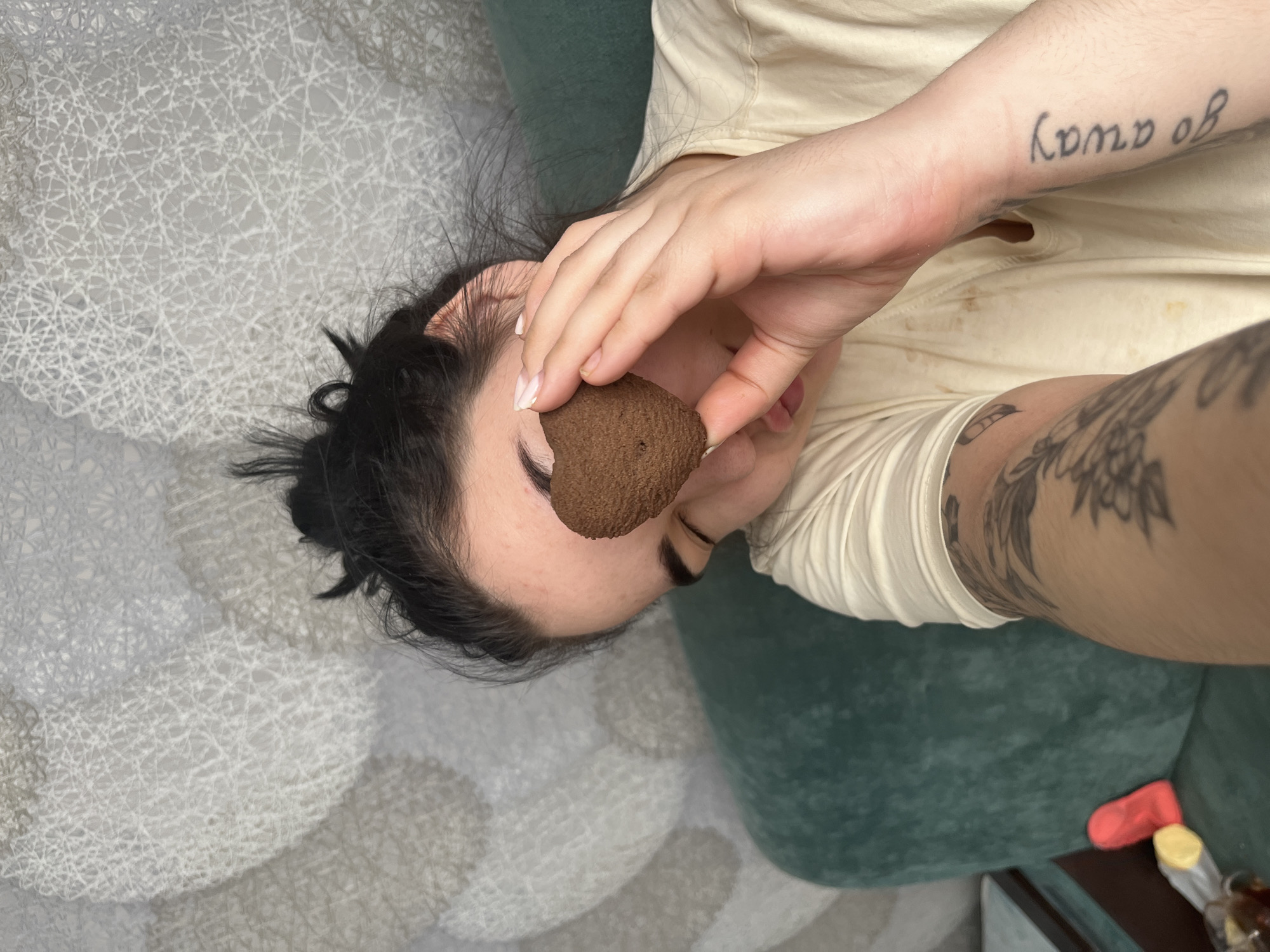 A woman is lying down, holding a heart-shaped, brown object in front of her face, obscuring her right eye. She has dark hair and light skin. Tattoos are visible on her left arm and wrist. She is wearing a light-colored shirt. She is lying on a green couch, which is situated on a patterned, gray and white rug. A red object and a bottle are partially visible on the side. No information related to Chaikovsky, Russia is visible.