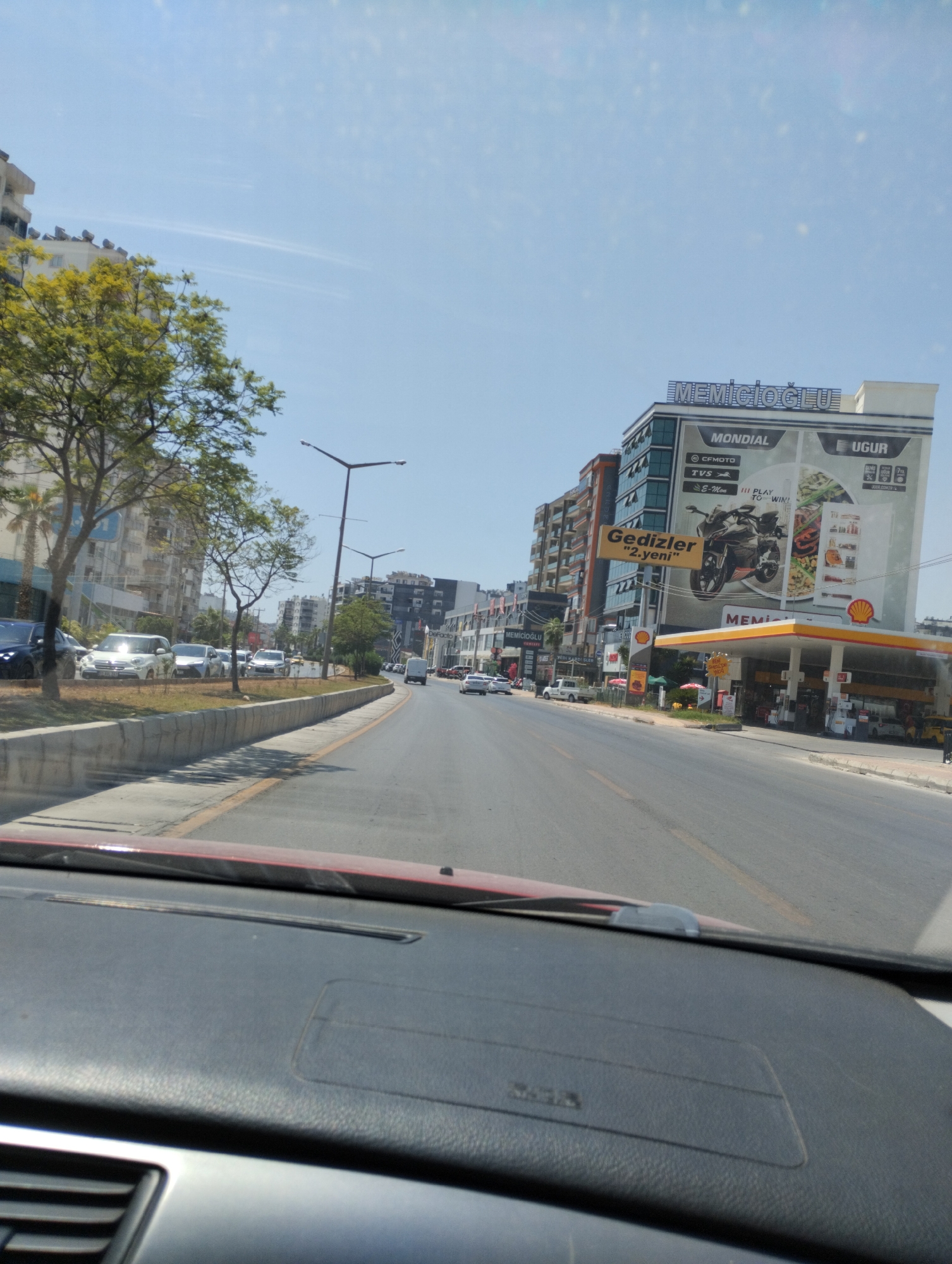 The image shows a street scene in Mezitli, Turkey, on a sunny day, viewed from the interior of a vehicle. The perspective is from the driver's seat, looking forward through the windshield. The road is paved with asphalt and has lane markings. On the right side of the road, a Shell gas station is visible with its yellow canopy. Above the gas station, a multi-story building features large advertisements, including "MEMİÇOĞLU" and "MONDİAL." To the left of the gas station, there are other buildings and trees lining the street. Several cars are parked and driving on the road. The foreground shows the dashboard and windshield frame of the vehicle. The sky is clear and blue with some wispy clouds.