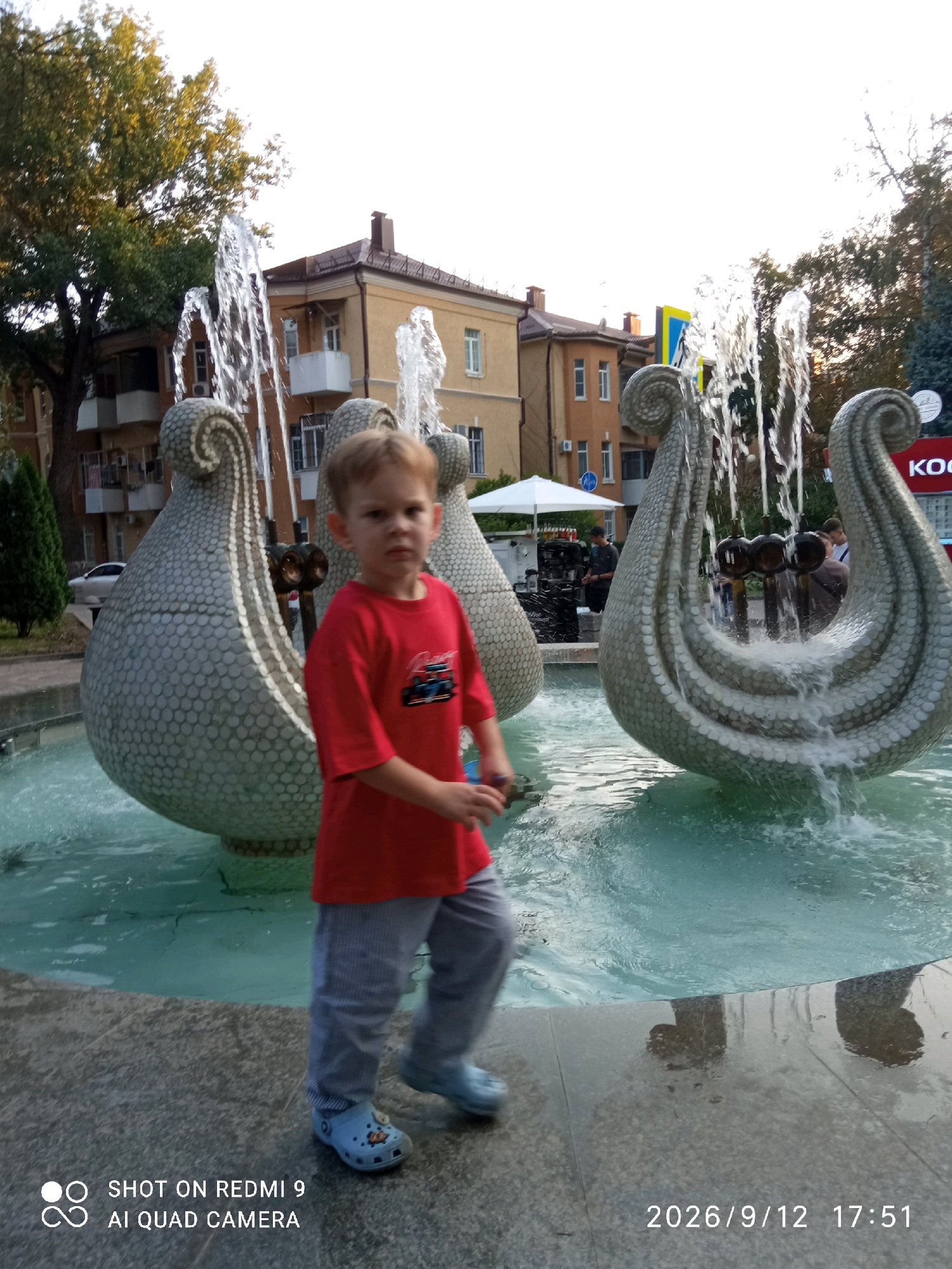 In the city of Rostov-on-Don, Russia, a young boy in a red t-shirt and grey striped pants walks past a decorative fountain. The fountain features large, light-colored, shell-shaped sculptures from which water sprays. In the background, there are several buildings with brown roofs and light-colored facades, as well as trees. The sky is bright, suggesting daytime. The image also contains a timestamp and text indicating it was taken with a Redmi 9 AI Quad Camera.