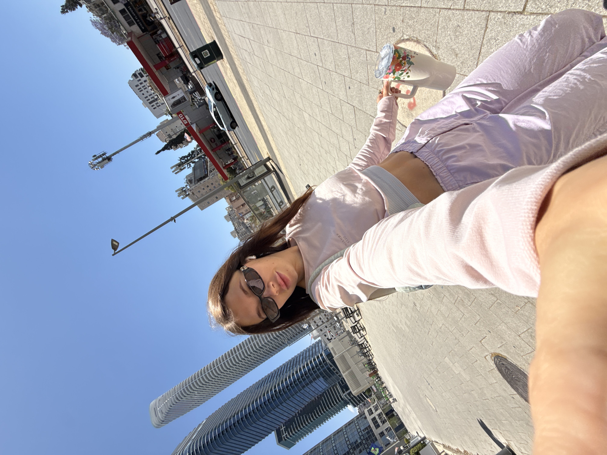 A young woman is taking a selfie on a sidewalk in Ramat Gan, Israel. The background includes the cityscape, featuring modern high-rise buildings. A gas station and other urban infrastructure are also visible. The woman is wearing sunglasses, a pink jacket, and lilac pants. She is holding a cup with a floral design. The photo is taken outdoors on a sunny day.