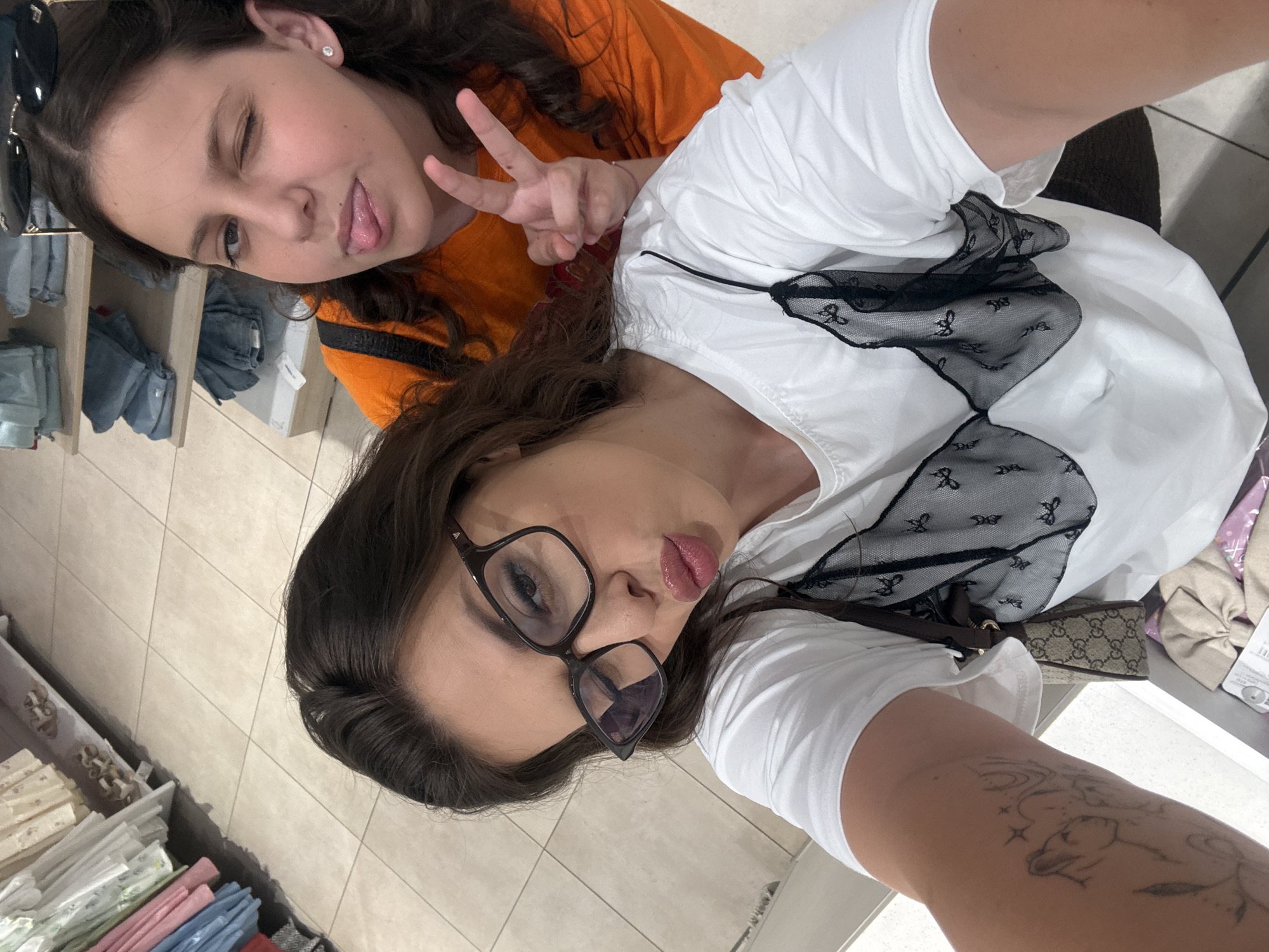 Two young women are taking a selfie in what appears to be a retail environment. The woman in the foreground, wearing glasses and a white t-shirt with a black lace overlay, is puckering her lips. The woman behind her, wearing an orange t-shirt, is winking and sticking out her tongue. The background shows shelves stocked with clothing and shoes. The floor is tiled. The scene is likely an indoor shopping area. There is no information in the image to indicate the location is specifically in Plovdiv, Bulgaria.