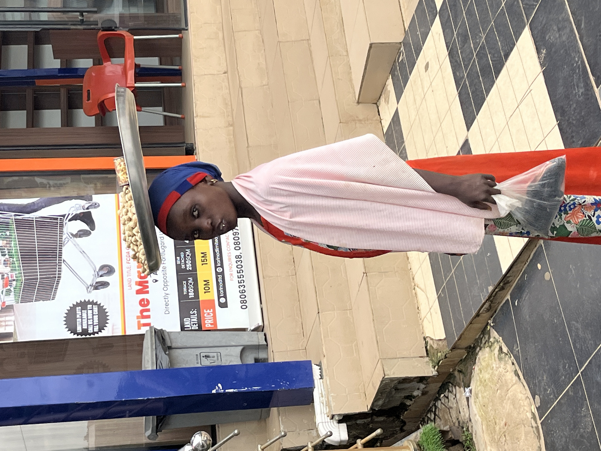 In Galadimawa Village, Nigeria, a young girl stands outside a building with a blue and metallic facade. She wears a blue headscarf with a red trim and a pink shawl draped over a red and floral-patterned dress. The girl balances a metal tray filled with food items on her head and holds a plastic bag containing dark material in her right hand. In the background, a red plastic chair is visible next to a shelf unit, and a sign with text and images related to land sales is displayed. The ground is paved with a checkered pattern of light and dark tiles.