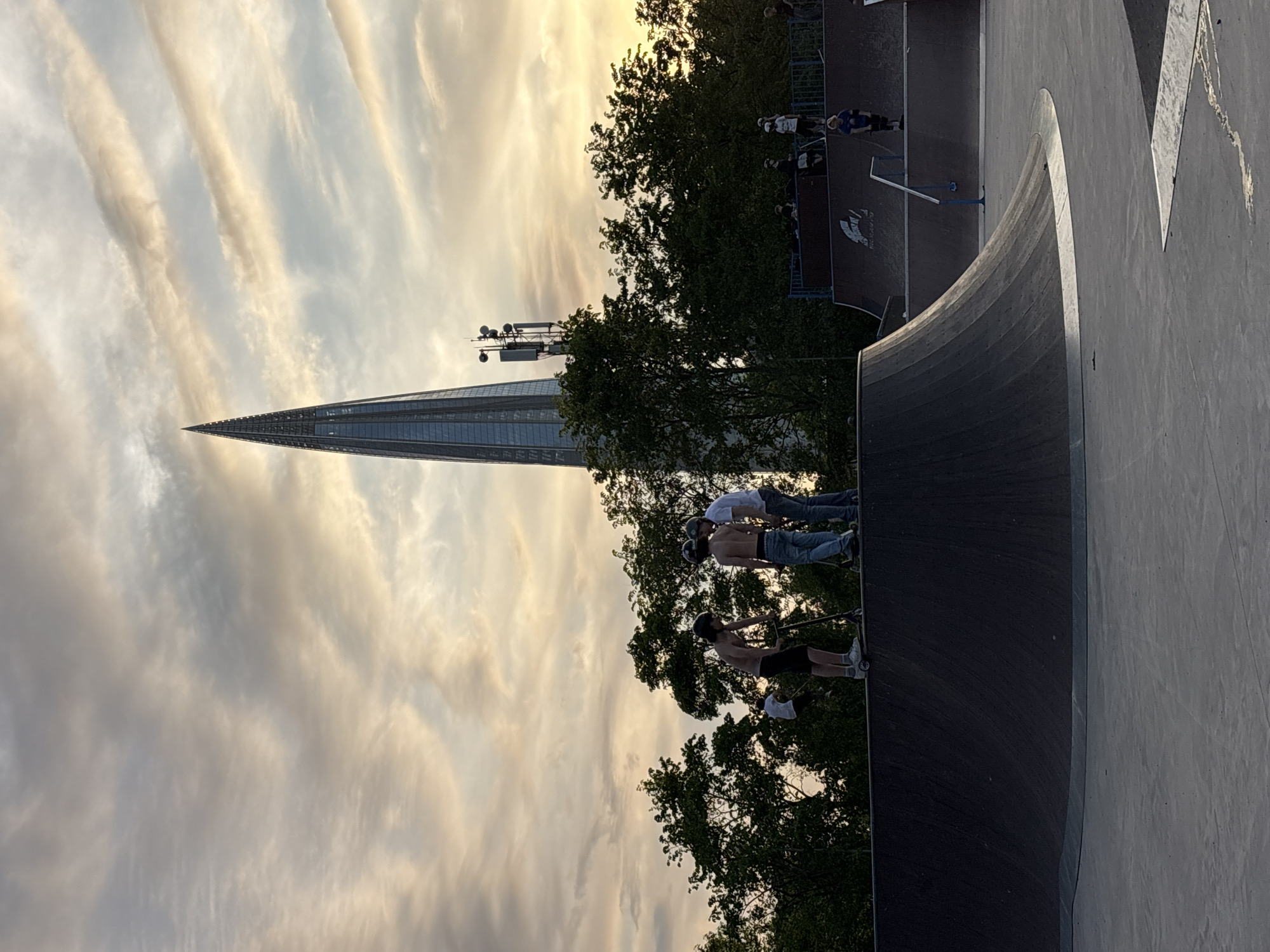 The image captures a dynamic scene at a skate park during the golden hour or late afternoon, characterized by warm, soft light. The sky is a prominent feature, displaying elongated, wispy clouds bathed in the sun's glow.
In the background, a tall, modern skyscraper with a distinctive pointed apex, resembling the Lakhta Center, rises prominently on the left side, towering above a canopy of lush green trees. A cellular tower or antenna array is also visible near the skyscraper, just above the treetops.
The foreground and midground are dominated by the skate park. A large, dark quarter pipe or ramp curves into the right side of the frame, its surface reflecting the warm light. On the edge of this ramp, three young men are present; two are shirtless, wearing shorts and jeans respectively, while the third wears a white t-shirt. They appear to be resting or observing. Helmets are visible near them, and one man holds a scooter.
Further back, another skate park structure features railings and a platform. More individuals are seen here, some on scooters or skateboards, indicating an active recreational environment. A specific logo or text reading "ROWDY" with an accompanying wing graphic is visible on the surface of this structure.
The overall scene conveys a relaxed and casual atmosphere, capturing an urban leisure activity. The setting is consistent with a skate park in Saint Petersburg, Russia.
