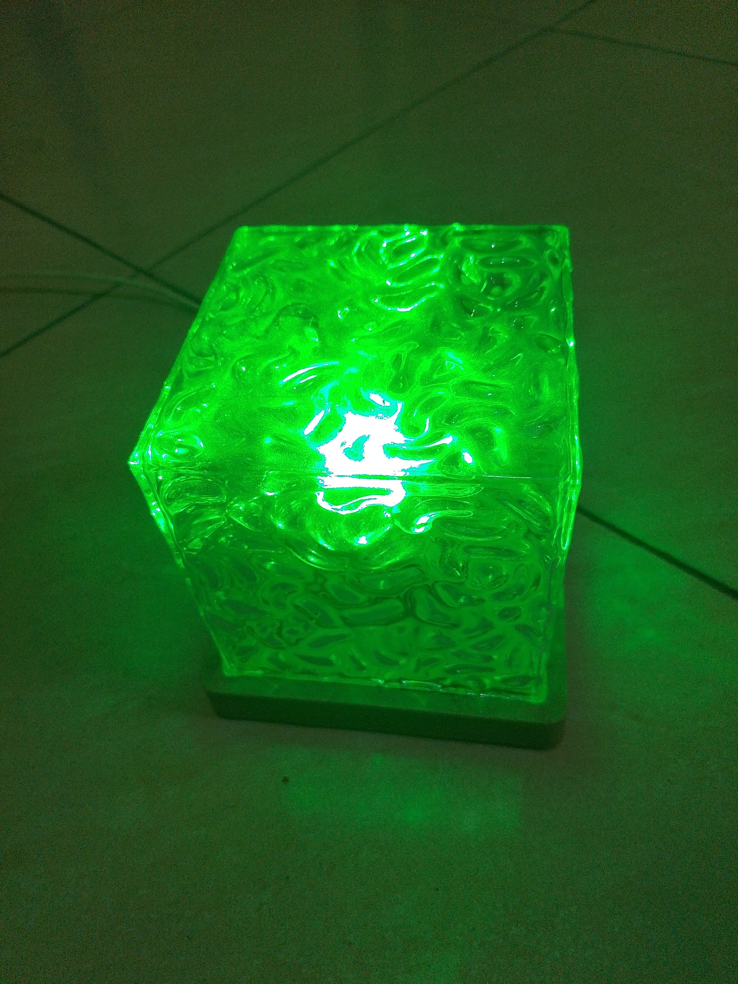 A close-up, overhead shot shows a translucent, green, cube-shaped object with a textured surface. The object emits a bright green light from within, illuminating the surrounding dark green, textured surface it rests upon. There are subtle, diagonal lines visible on the surface behind the object, possibly indicating wires or seams. The overall lighting is dim and casts a green glow across the scene. The location in the city of Ketanggung, Indonesia, is not discernible from the image.