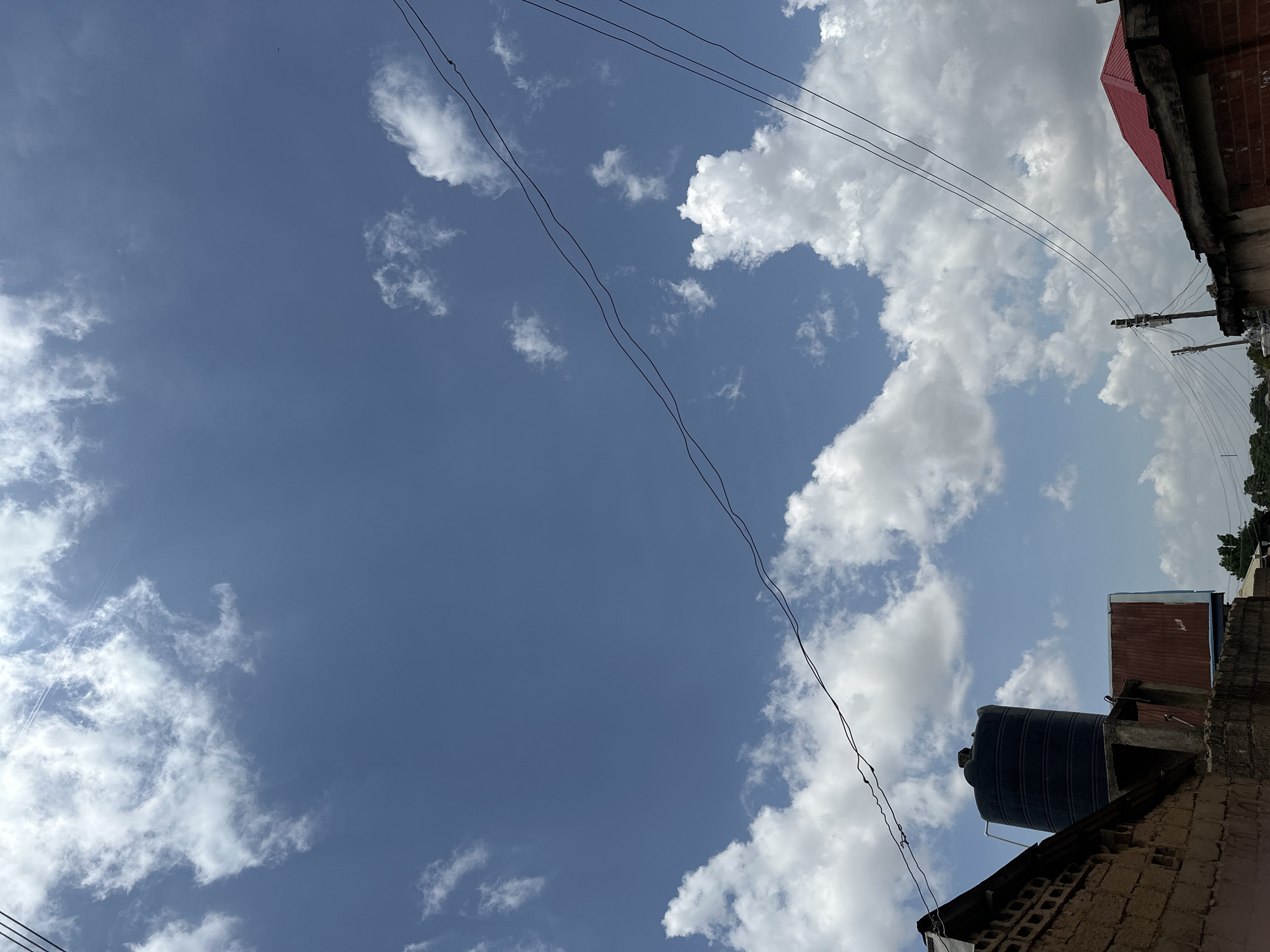 The image shows a daytime outdoor scene with a bright blue sky filled with scattered white clouds. In the lower portion of the image, various urban elements are visible. To the left, a brick wall is prominent, with a black cylindrical water tank on top. Next to it, a building with a rusted metal roof is partially visible. Several power lines stretch across the frame, intersecting the sky. In the lower right, another building with a red corrugated metal roof is seen. A utility pole with wires is also present. The overall environment appears to be a residential or mixed-use area in a city.