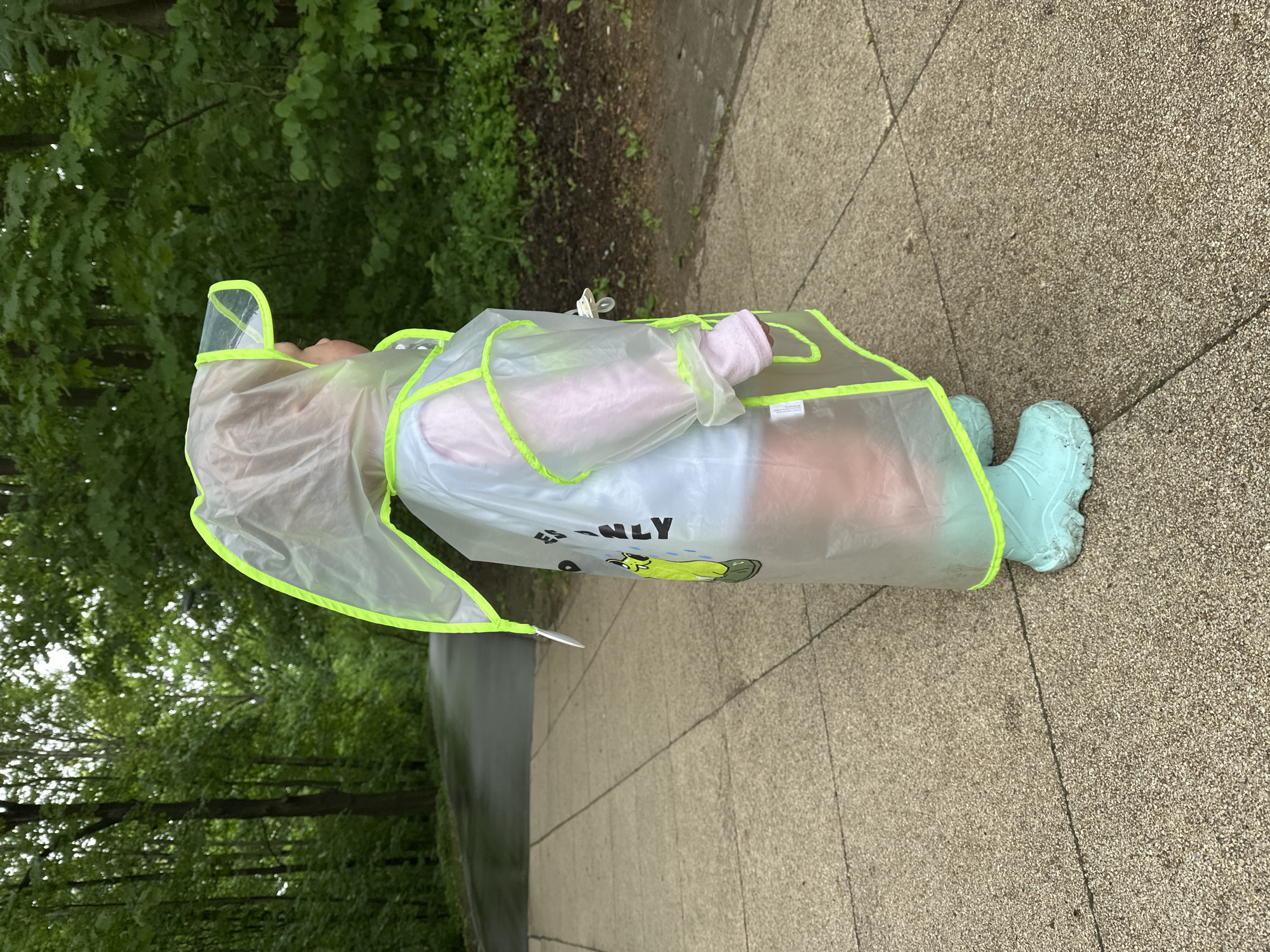 A child wearing a clear raincoat with neon green trim and light blue rubber boots stands on a light gray concrete sidewalk. The raincoat has a clear visor and a cartoon character on the front. The child's hand is covered with a light pink sleeve. The background features green trees and foliage, suggesting a park or green space. The location in Nizhny Novgorod, Russia, cannot be confirmed from the image alone.