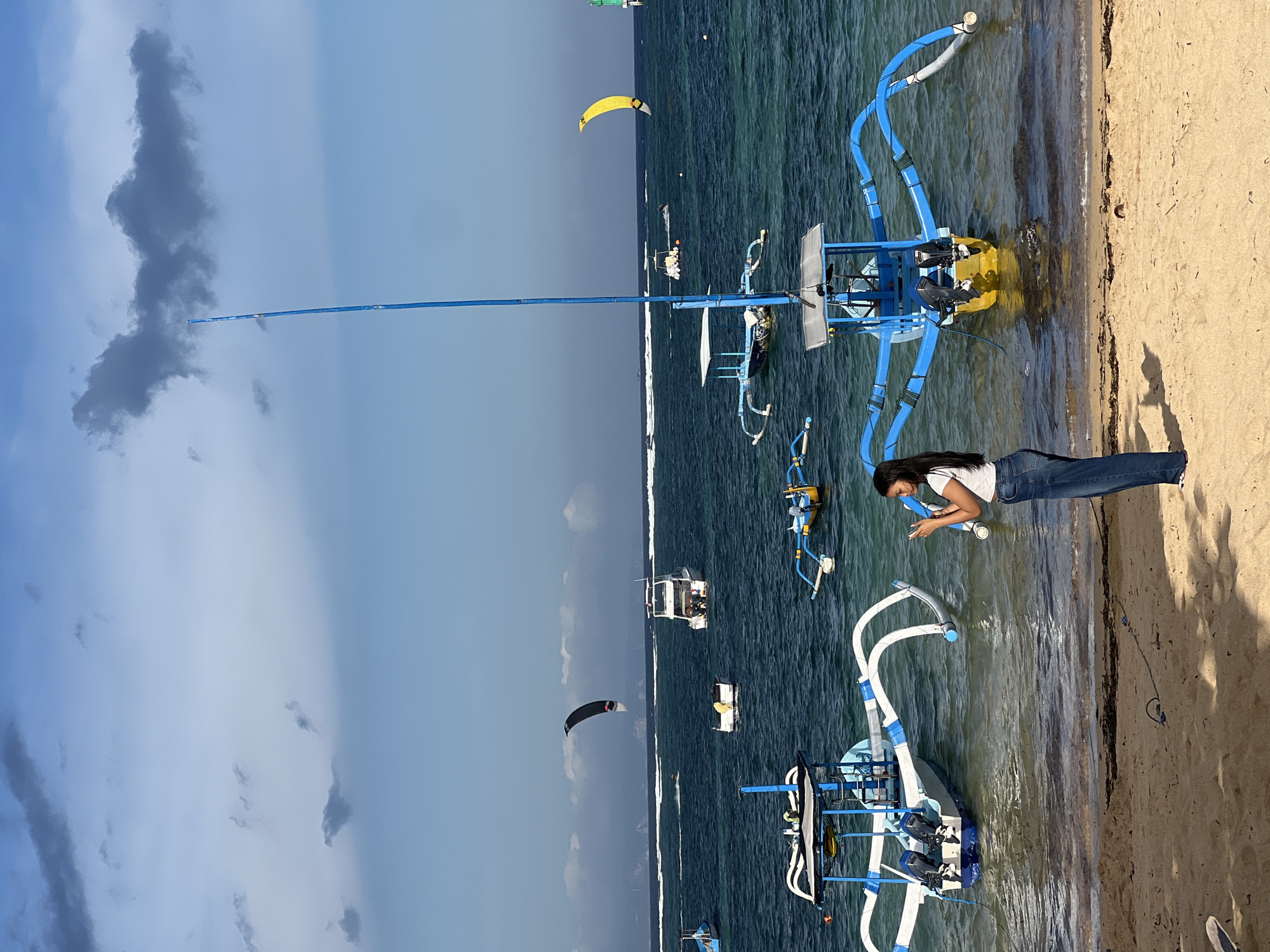 This is a vertical, outdoor, daytime photograph taken in Denpasar, Indonesia. The scene depicts a beach with a person and several boats on the water. The sky is partly cloudy with some dark clouds, suggesting a dynamic weather condition, possibly transitioning from rain or preparing for it. The sea is a deep blue-green, indicating clear but potentially rough waters, with waves breaking near the shore.
In the foreground, a person with long dark hair is leaning on a blue and yellow boat, looking at their phone. They are wearing a white t-shirt and blue jeans. The boat is a traditional outrigger canoe with a distinctive blue frame structure. Further out in the water, several other similar boats are visible, some with people in them.
In the distance, there are indications of water sports activity. A yellow kite is visible in the sky, and further to the left, a black and white kite is also in the air, suggesting kite surfing or kiteboarding. The presence of these kites indicates that there are individuals engaged in these activities on the water.
The beach itself is sandy, with some seaweed and debris along the waterline. The sun appears to be casting shadows, but the overcast sky might be diffusing the light. The overall mood of the image is one of leisure and outdoor activity, with a hint of the power of nature due to the cloud formations and sea conditions. No visible text is present in the image.