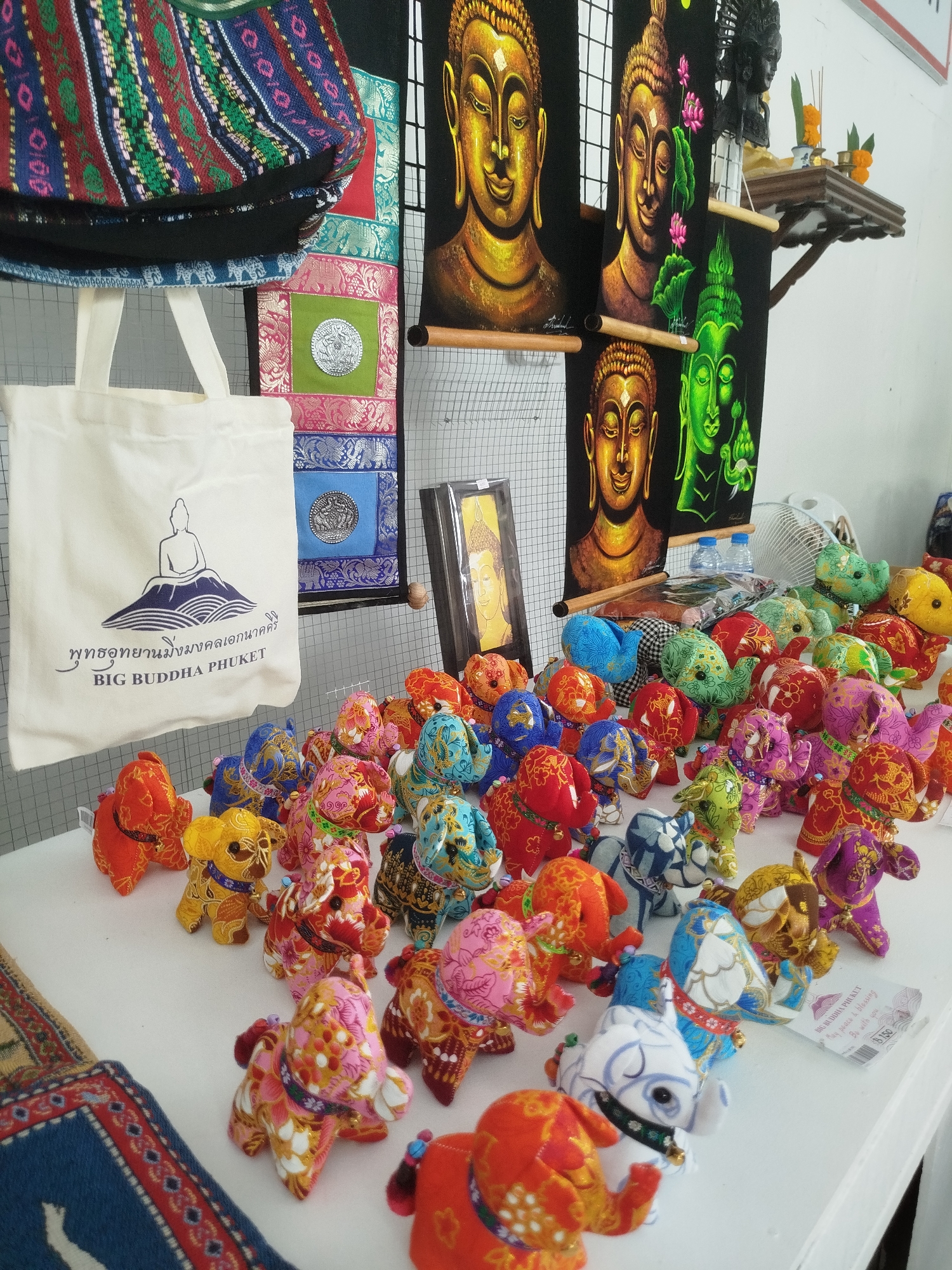 This media file showcases a vibrant stall likely selling souvenirs in Karon, Thailand. The foreground is dominated by a collection of colorful, fabric-stuffed elephants, each adorned with intricate, traditional patterns. These charming trinkets are arranged in a dense cluster on a white surface, suggesting a market or shop setting.
Behind the elephants, a large, off-white tote bag with a printed image of a meditating figure on a mountain and Thai text is prominently displayed. The bag also features the English text "BIG BUDDHA PHUKET," providing a clear location cue.
Further in the background, several black fabric tapestries hang, each featuring a striking image of a Buddha's face. Some are rendered in gold, while another is depicted in vibrant green. These are accented with lotus flowers and leaves. A small, wooden shelf above the tapestries holds an assortment of Buddhist offerings, including incense sticks and small orange flowers, reinforcing the cultural context. A white electric fan is partially visible in the far right background, suggesting an indoor or semi-outdoor stall.
The overall impression is one of a lively, culturally rich marketplace, likely a tourist-oriented shop due to the souvenir items and the prominent "BIG BUDDHA PHUKET" branding. The bright colors and the variety of items create a visually appealing and inviting scene. A small price tag with the text "B150" is visible near the bottom right, indicating the cost of one of the elephant souvenirs.