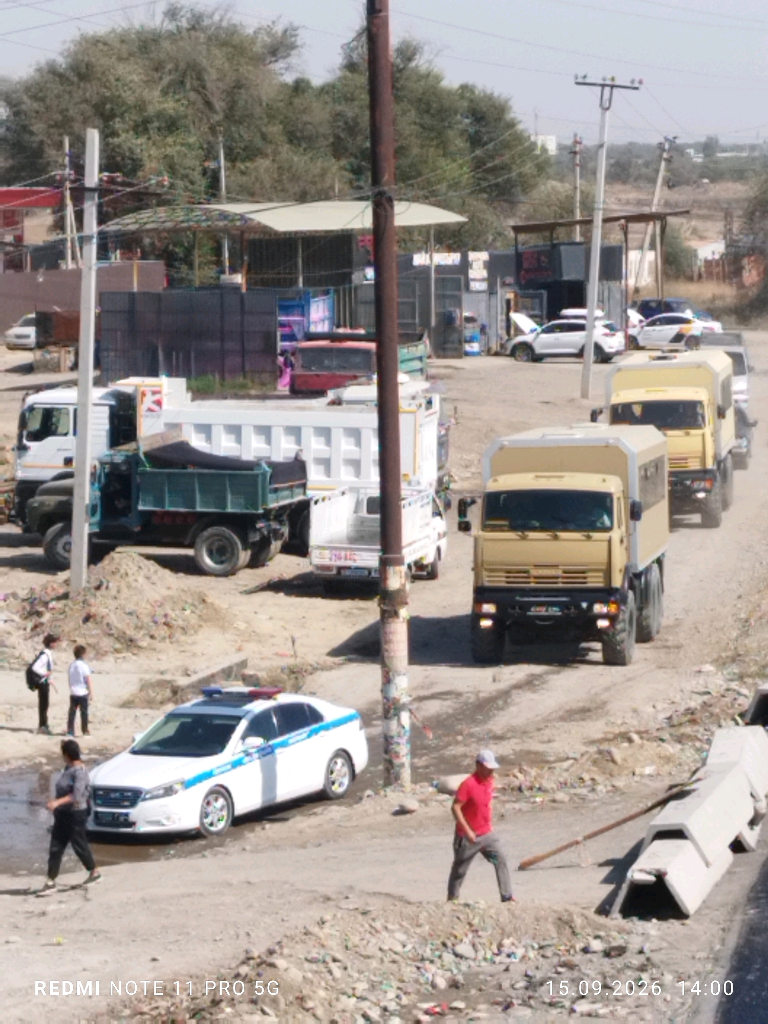 The image depicts an outdoor scene in a dusty, semi-developed area, likely on the outskirts of Bishkek, Kyrgyzstan. In the foreground, a white police car with blue and red lights on its roof and blue stripes along its side is parked. Two individuals stand near the police car, one a male in dark clothing and the other a male in a white shirt and dark pants. Further back, a convoy of beige trucks is proceeding down a dirt road. The lead truck is a six-wheeled vehicle. Behind it, two similar trucks are visible, with one slightly obscured. To the left of the police car, a white dump truck with a green bed is parked, and to its left, another white truck is visible. In the background, buildings and trees can be seen, suggesting an urban or peri-urban environment. The ground is covered in dirt, gravel, and some scattered debris. A concrete barrier is visible on the right side of the image. The overall impression is of an organized movement or operation involving the police and military or transport vehicles.
