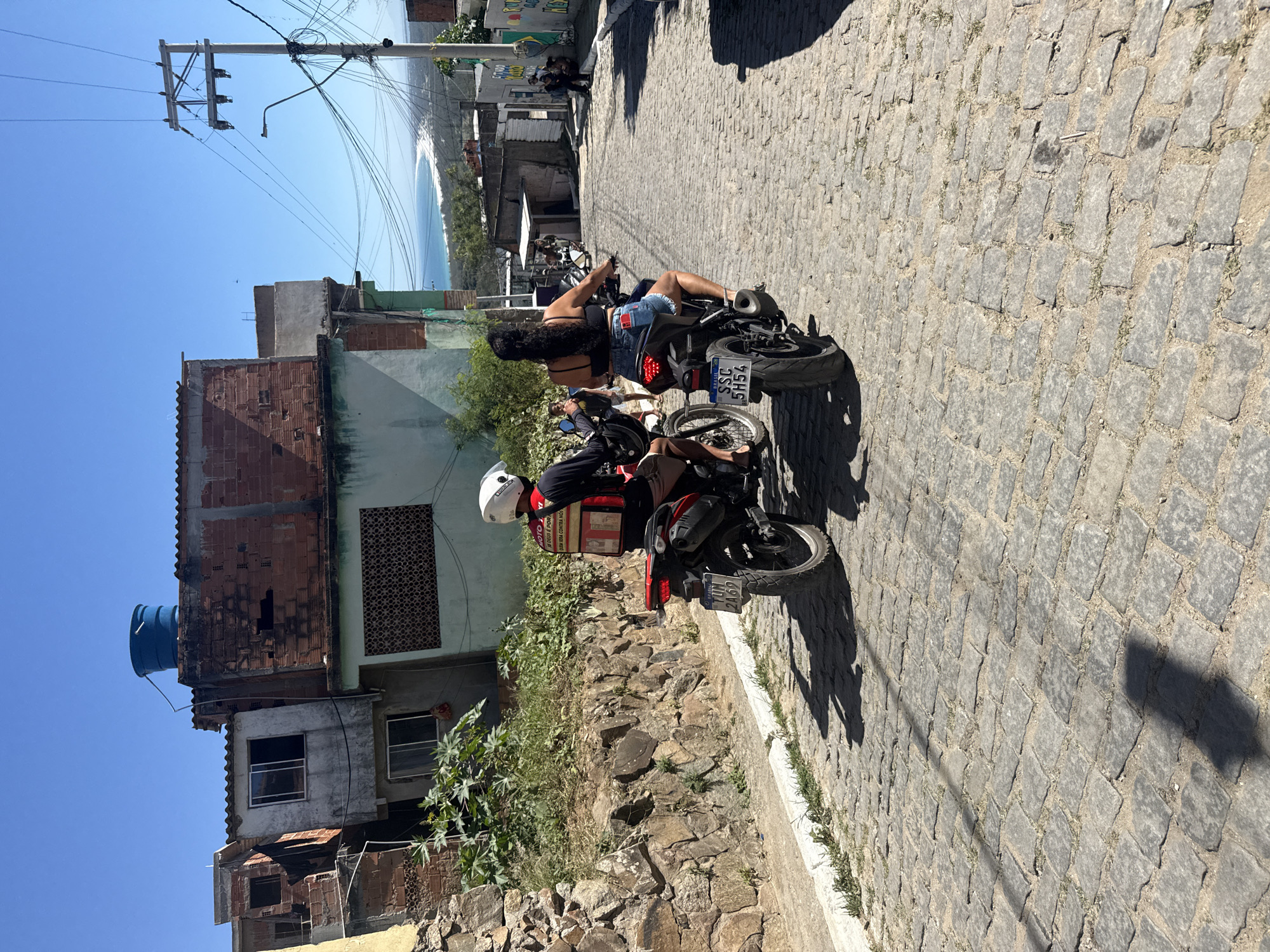 In Arraial Do Cabo, Brazil, two people ride motorcycles up a cobblestone street under a clear blue sky. The person in front, wearing a white helmet and a red vest with white lettering, is on a red motorcycle. The person behind, wearing a black top and denim shorts, is on a black motorcycle with a license plate that reads "SSC 5H54". The street is lined with stone walls and houses, some appearing unfinished. In the background, the ocean and a beach are visible.