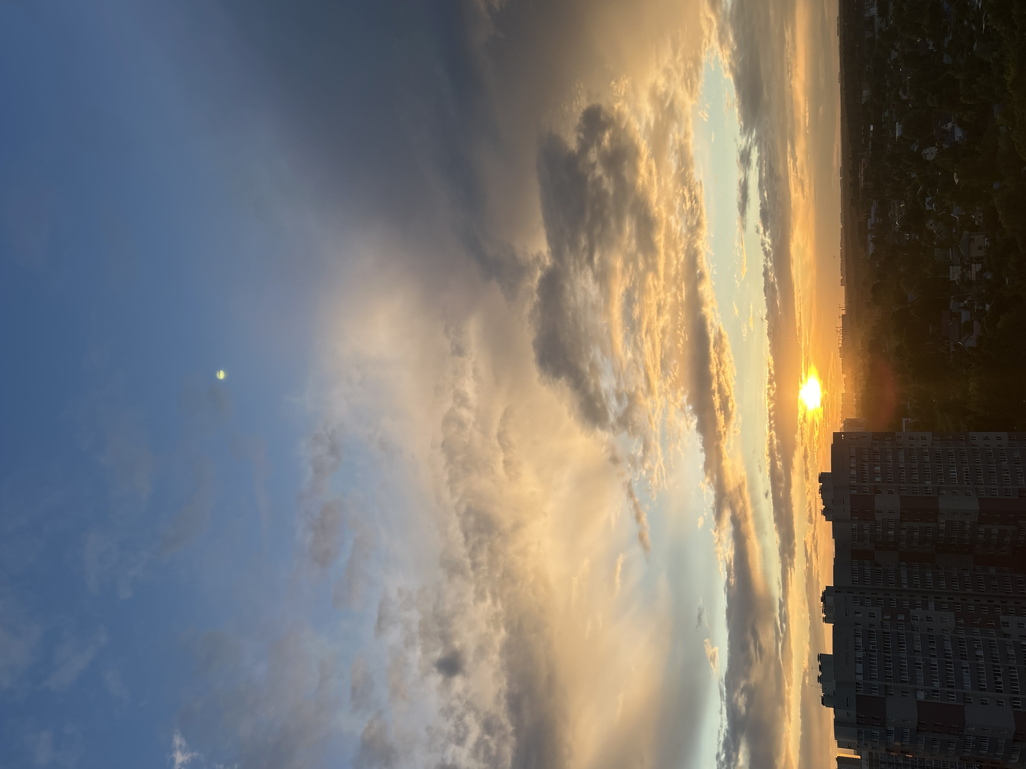 The image shows a wide-angle view of a city skyline at sunset. A large, multi-story apartment building dominates the left side of the frame, with a dense residential area and trees stretching into the distance on the right. The sky is filled with dramatic clouds illuminated by the setting sun, which is a bright orange orb visible near the horizon. A small, pale yellow circle, possibly the moon or a lens flare, is visible higher up in the blue sky. The overall impression is of a peaceful, urban landscape bathed in the warm glow of twilight. The location is identified as Ivanteyevka, Russia, based on contextual information.