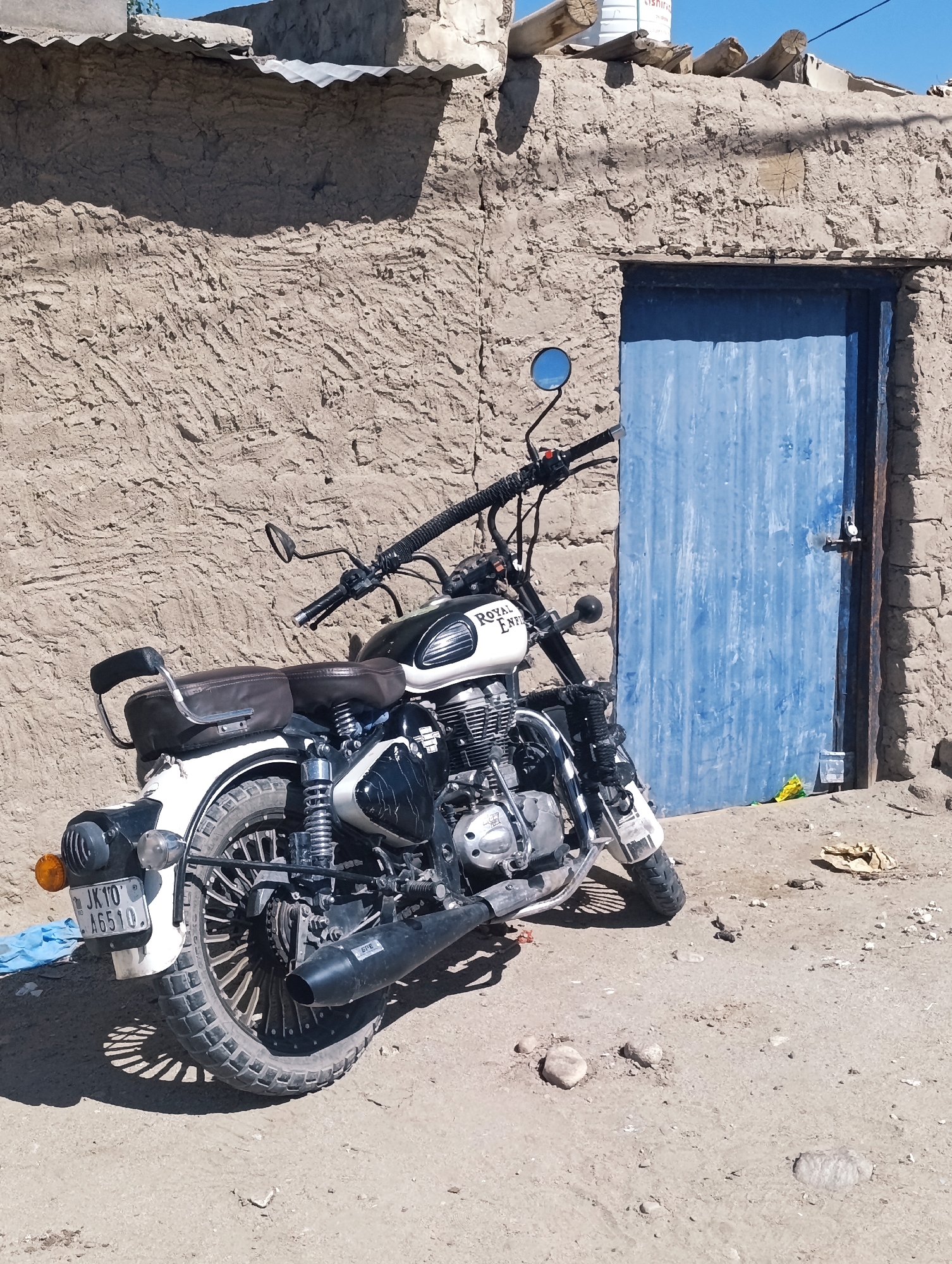 In the city of Leh, India, a black and white Royal Enfield motorcycle is parked in front of a rustic, mud-brick building. The building features a corrugated metal roof and a blue wooden door with a visible lock. The motorcycle's license plate is partially obscured but displays the characters "JK 10 A6510". The ground is dusty and uneven, with scattered stones and debris. The scene appears to be outdoors, under a clear blue sky.