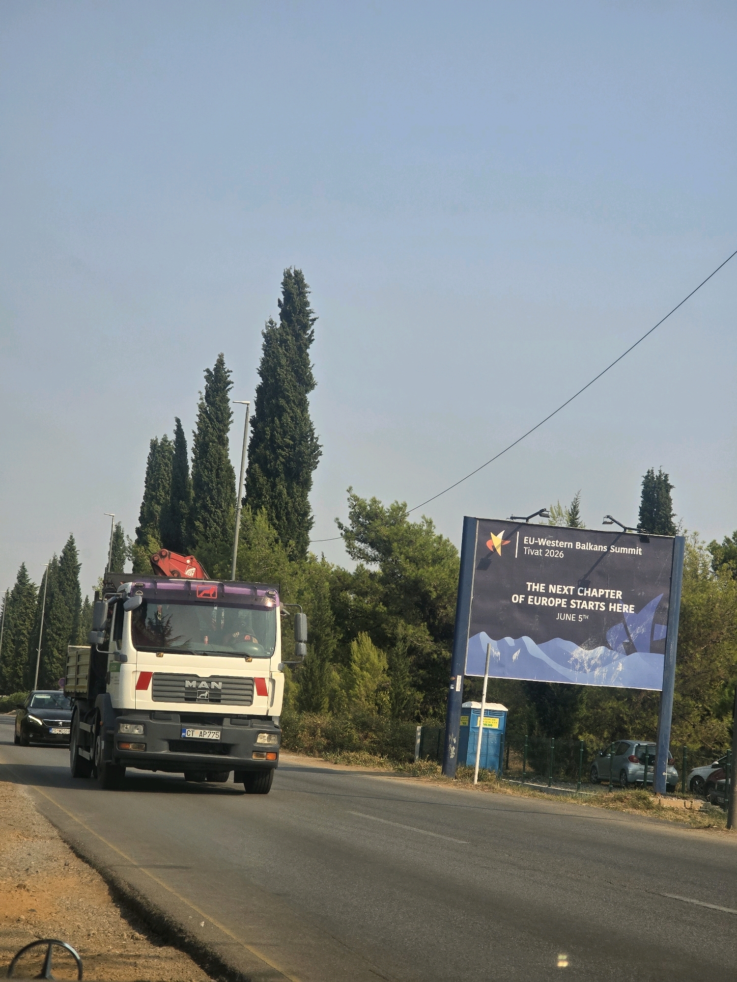 This is a medium-angle, eye-level shot of a road in Mojanovići, Montenegro, on a clear, sunny day. The road is paved, with a dirt shoulder on the left. In the foreground, a white truck with a red crane is driving on the road. The truck has the brand "MAN" visible on its front. A black car is driving behind the truck. To the right of the road, there is a large billboard advertising the "EU-Western Balkans Summit Tivat 2026." The billboard states, "THE NEXT CHAPTER OF EUROPE STARTS HERE JUNE 5TH." A blue portable toilet is located next to the billboard. In the background, there are many tall, green trees and a clear blue sky. Several streetlights are visible amongst the trees.