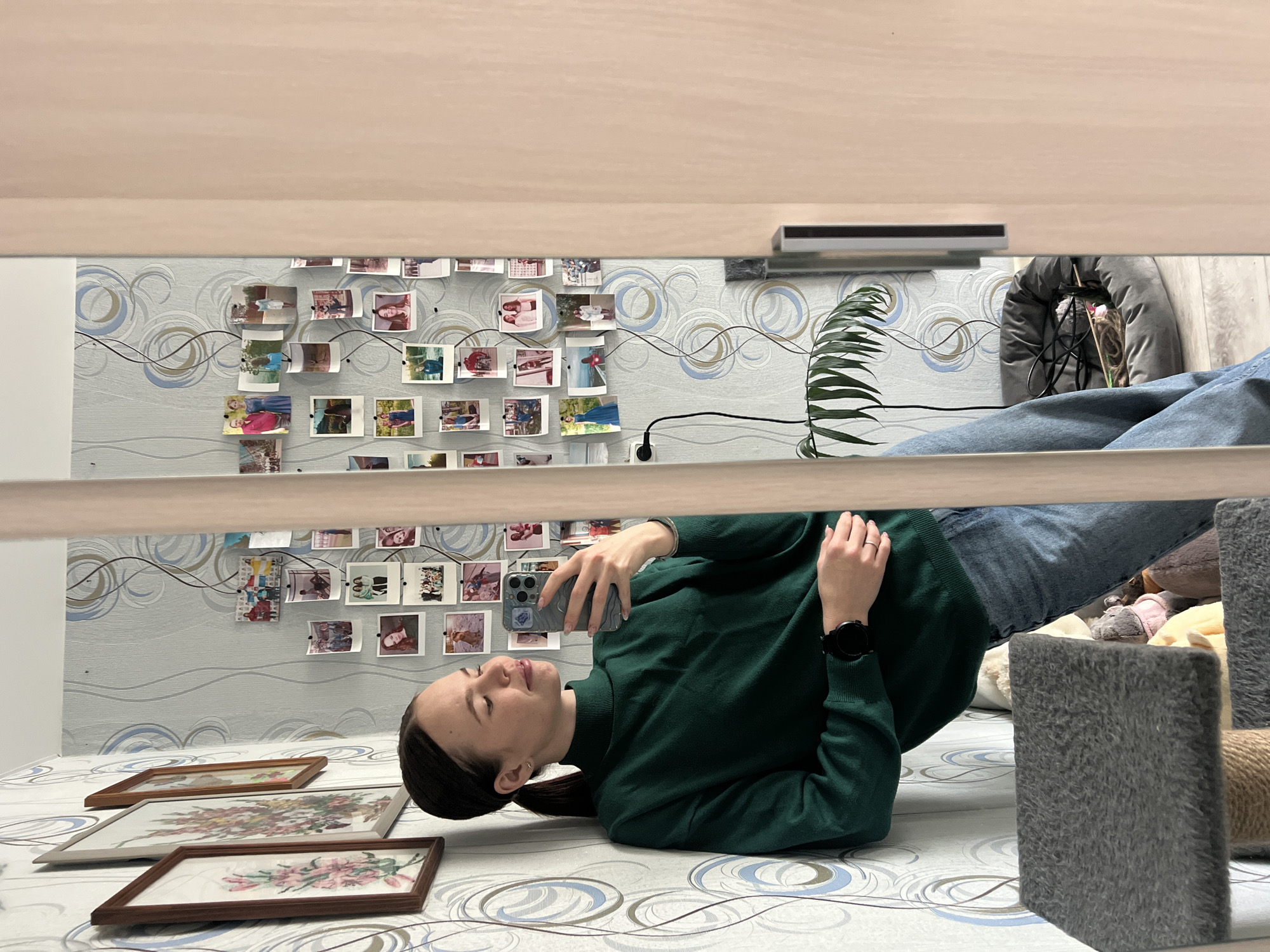 A woman lies on a bed in a room, holding a smartphone. The room has wallpaper with a pattern of circles and lines, and a display of photographs on the wall. There are framed pictures on the bed. A plant and a pet bed are visible in the background. Part of what appears to be a desk or shelf is above the woman. While the location is suggested as Chaikovsky, Russia, there are no verifiable details in the photo to confirm the specific location.