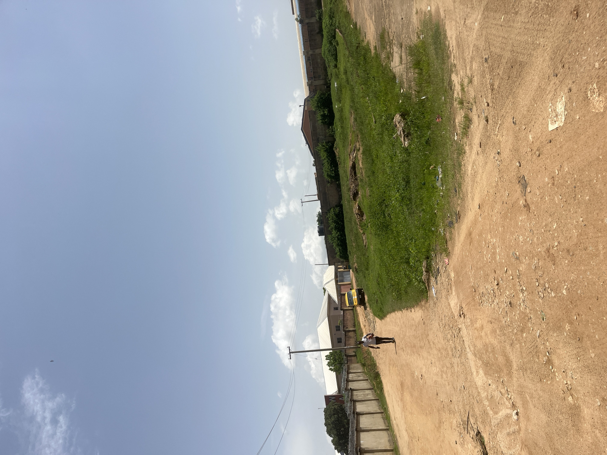 A man is walking away from the camera on a dirt road in Gura Top, Nigeria. He is wearing a dark t-shirt and light-colored shorts. A yellow school bus is parked on the right side of the road, near some buildings and a concrete wall with barbed wire. The area is dry and dusty, with patches of green grass and some scattered debris. The sky is clear with a few scattered clouds.