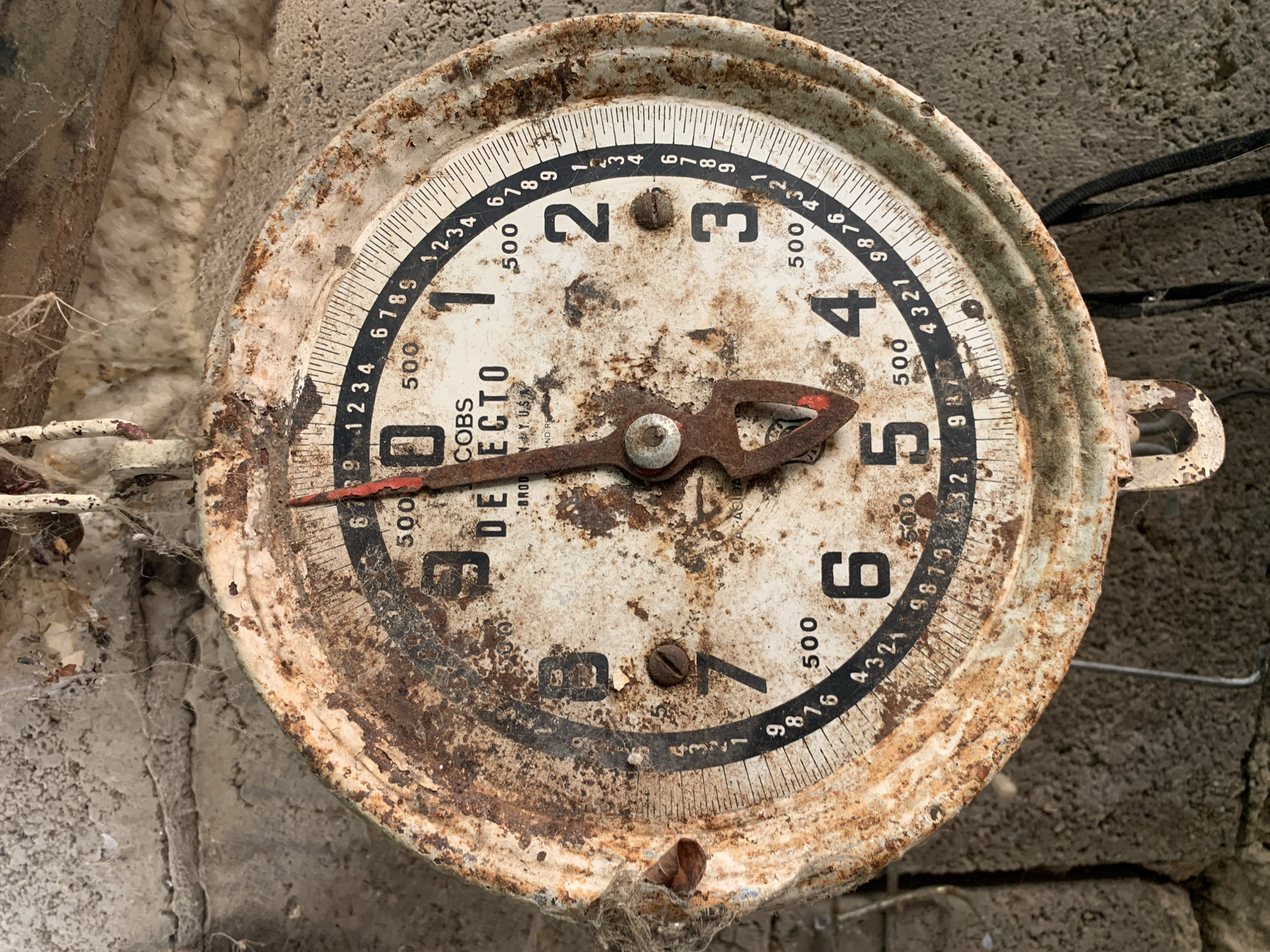 This is a close-up, forensic, matter-of-fact image of an old, heavily rusted, circular scale. The scale has a white face with black numbers and markings, heavily obscured by rust and dirt. A red-tipped pointer is positioned just past the 12 on the scale. The numbers visible are 1, 2, 3, 4, 5, 6, 7, 8, and 9, with smaller increments marked between them. The words "COBS DEJECTO BROOKLYN N.Y. U.S.A." are visible on the face of the scale. The scale is hanging from a rusted hook against a rough, textured, gray concrete wall, with visible cobwebs and insulation material in the background. The image does not contain any information that would allow for the identification of its location within the city of Santa Ana, Venezuela. There are no people, actions, or interactions depicted in the image.