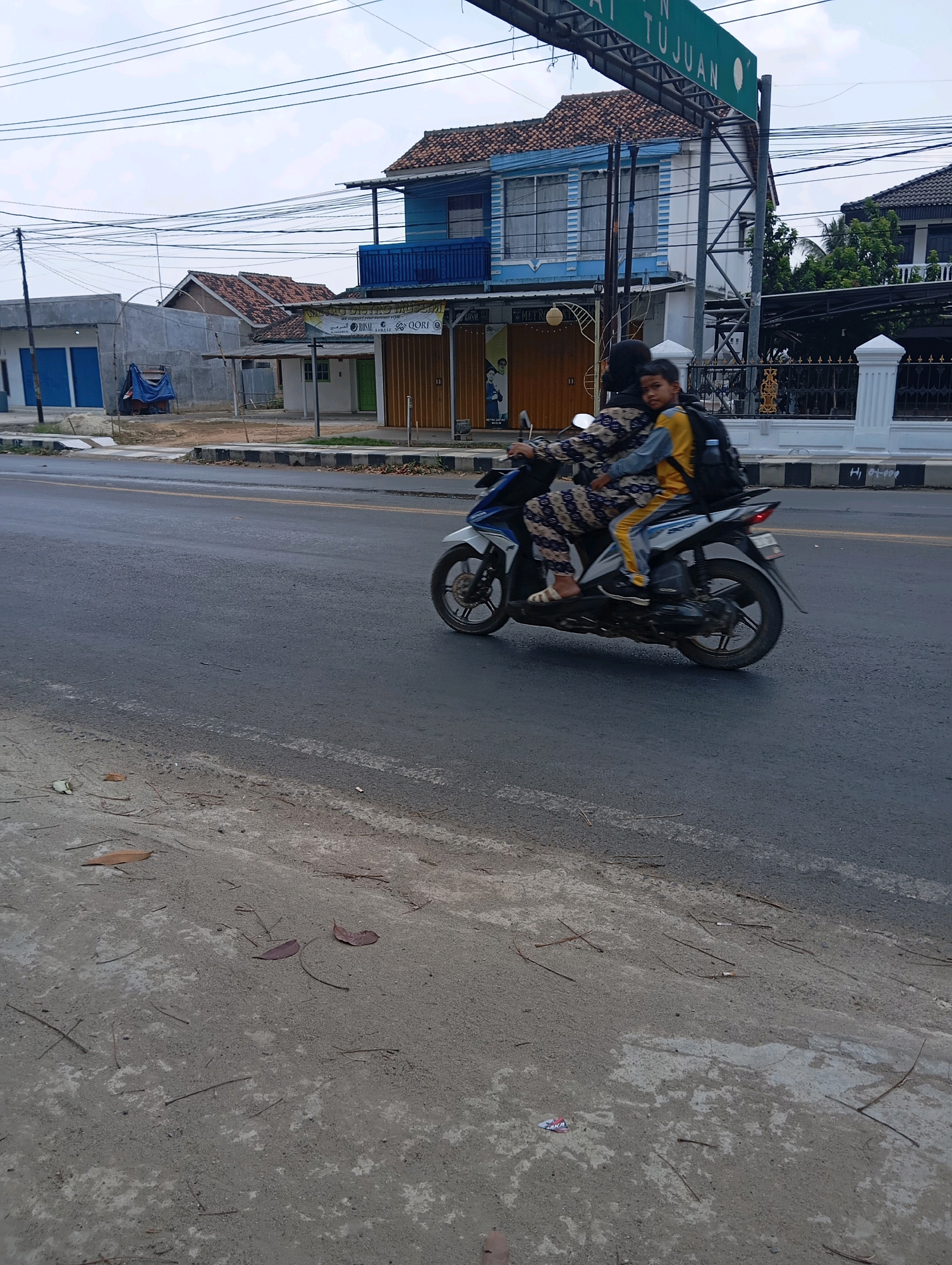 In Metro, Indonesia, a person wearing a patterned outfit and a headscarf rides as a passenger on a motorbike. They are seated behind the driver, who is not fully visible. The passenger has their arms around the driver and is looking forward. They are wearing a yellow and gray jacket and dark pants, and have a backpack on. The motorbike is moving on a paved road. In the background, there are buildings, including a blue and white building with a balcony, and a sign that reads "TUJUAN" (meaning "Destination"). The sky is cloudy. The foreground shows a dirt and gravel shoulder of the road with some fallen leaves and twigs.