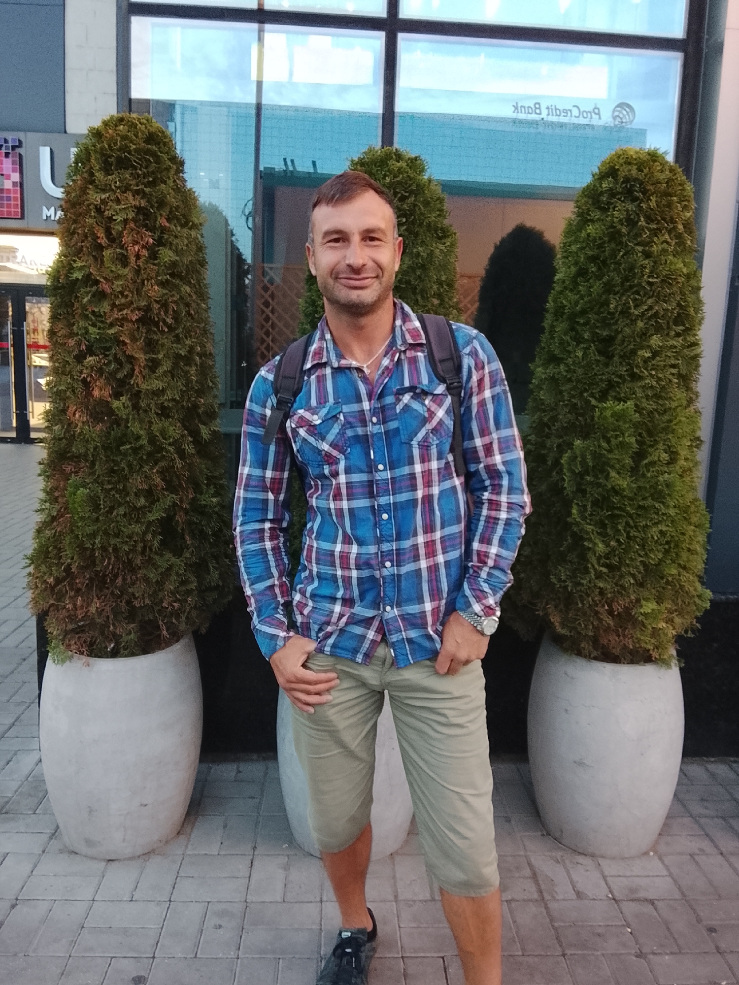 A man stands outdoors between two tall, conical evergreen trees in large, light-colored planters. He is wearing a blue and red plaid shirt, light tan shorts, a silver wristwatch, and dark sneakers. He has his left hand in his pocket and his right hand is by his side. The background features a modern building with large windows, some of which reflect the sky. A sign in one of the windows reads "ProCredit Bank." The ground is paved with gray rectangular bricks. This appears to be an urban setting. Based on the presence of "ProCredit Bank," a financial institution with a presence in Chișinău, Moldova, this scene is likely located in Chișinău, Moldova.