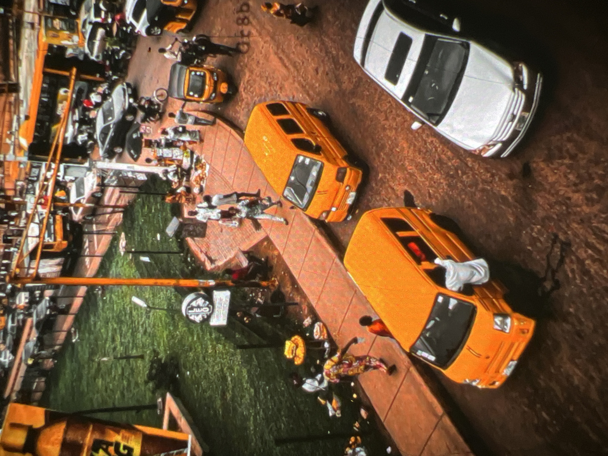 This aerial photograph captures a bustling street scene in Lagos, Nigeria. The image is dominated by various vehicles, including several orange minibuses, a white sedan, and a yellow motorized tricycle. The road appears to be paved with rough, reddish-brown material. On the left side of the frame, a grassy area is visible, featuring a large, yellow bottle-shaped advertisement and some people walking. A raised sidewalk, paved with orange tiles, runs alongside the grassy area and the road. Several people are seen on the sidewalk and on the road, engaged in various activities. Some are walking, others are standing near vehicles, and a few appear to be vendors with goods displayed on a table. The overall impression is of a lively and active urban environment.