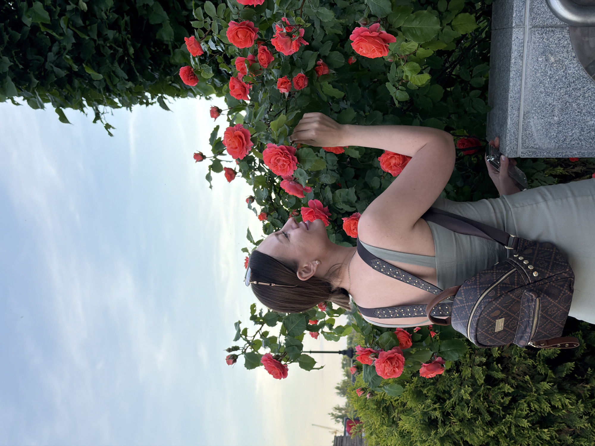 A woman is standing next to a rose bush with red roses. She has a brown backpack on her back and is wearing a green dress. The scene is taken outdoors with the sky visible in the background. The location is Rostov-on-Don, Russia.