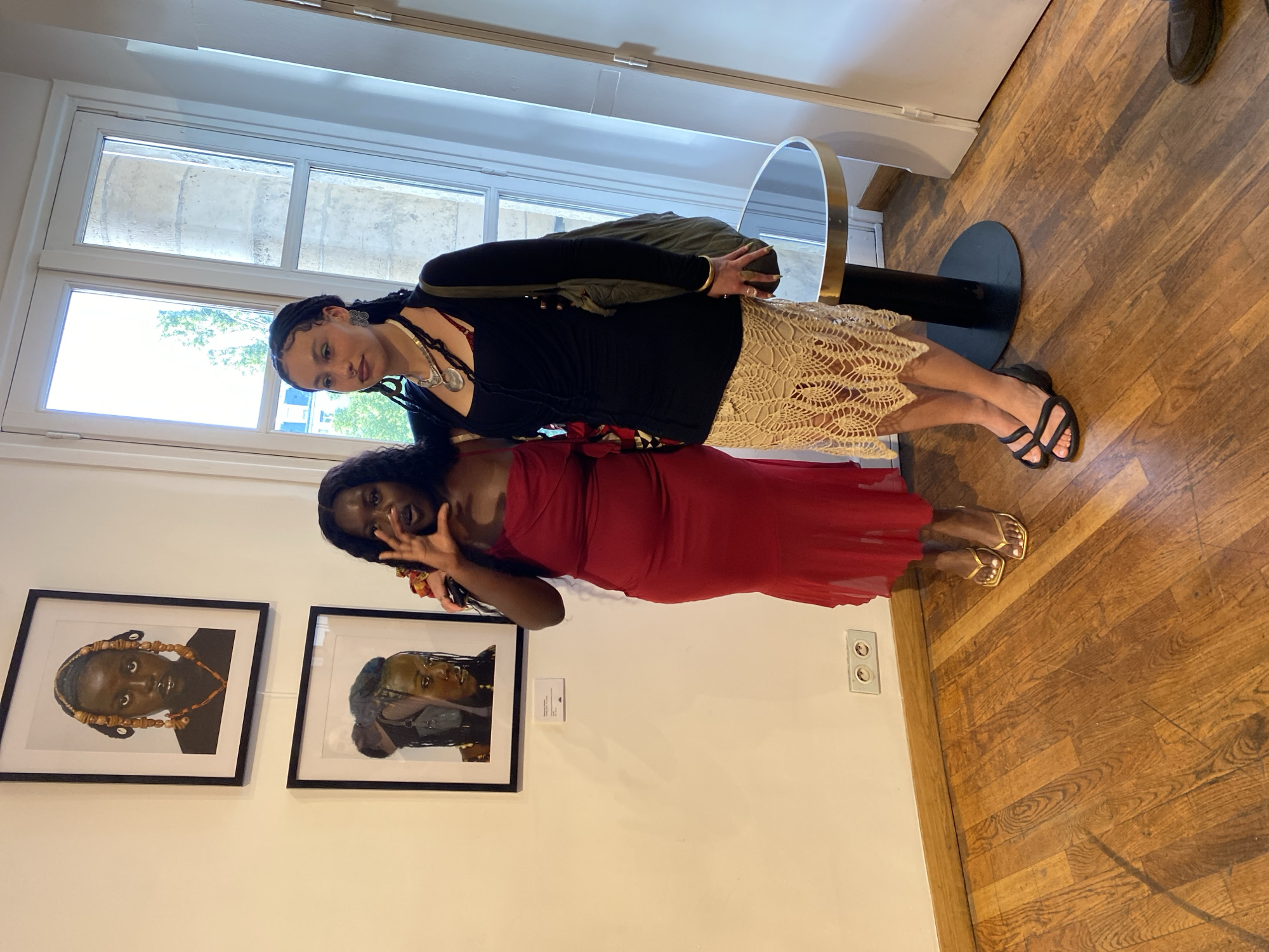 Two women pose in front of artwork displayed on a wall inside a building. The woman in the foreground wears a red dress and gold sandals. Behind her, the other woman is dressed in a black top, a cream-colored skirt, and black sandals. On the wall are two framed portraits of women. A window is visible in the background, showing buildings and sky outside. The floor is wooden. A round mirror is positioned near the window. The scene is set in Paris, France, as evidenced by architectural details.