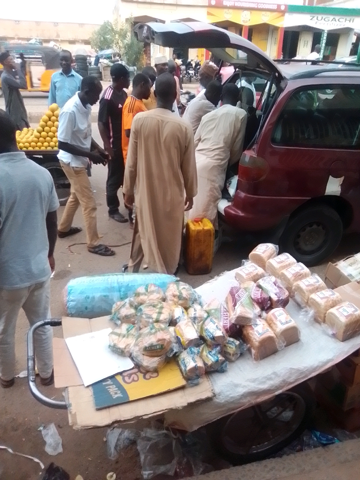 The image depicts a street scene in Arawa, Nigeria, focusing on a market stall laden with bread and other goods. Several people are present, some interacting with the stall and others in the background. In the foreground, a cart is visible with a large blue tarp and an abundance of packaged loaves of bread. The bread packaging features various brands and colors. Behind the cart, a maroon car with its rear hatch open is parked, with individuals appearing to load or unload items from it. A large yellow jerrycan is positioned on the ground next to the car. In the mid-ground, a man in a light blue shirt is seen near a cart filled with bright yellow corn or fruit. Further back, a yellow and black motorized tricycle is also visible. The environment appears to be an outdoor market setting with buildings and shops in the background, one displaying a sign that reads "ZUG ACHE FERTILIZER". The ground is paved with dirt and asphalt. The overall scene suggests a bustling commercial area, likely a street market in Arawa, Nigeria.