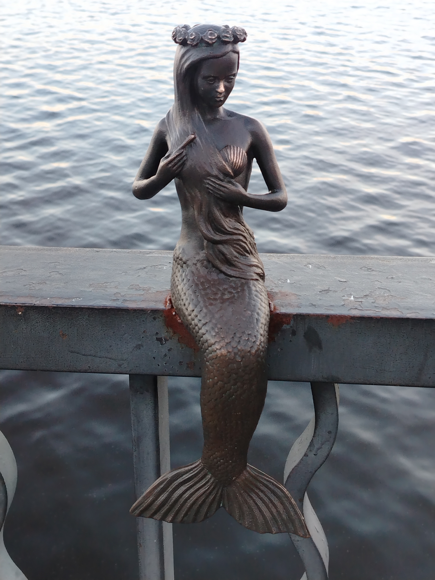 A bronze statue of a mermaid sits centrally on a dark metal railing, looking downwards with a contemplative or melancholic expression. She wears a floral wreath on her head, and her long hair flows over her left shoulder. Her hands are placed over her chest, with one hand covering a seashell-shaped bra and the other resting near her collarbone, possibly holding her hair or garment. The mermaid's body features intricate scales, and her tail ends in a fanned fin.
The statue is positioned on a bridge railing or waterfront balustrade, with a calm body of water, likely a river or lake, stretching into the background. The water's surface shows subtle ripples, reflecting a soft, diffused light, suggesting late afternoon or early evening. The metal railing supporting the mermaid exhibits signs of wear and rust, particularly where the statue makes contact. Below the main railing, decorative curved metalwork is visible. No discernible activity is taking place beyond the static presence of the sculpture. There is no visible text in the image. This scene is located in Chaikovsky, Russia.
