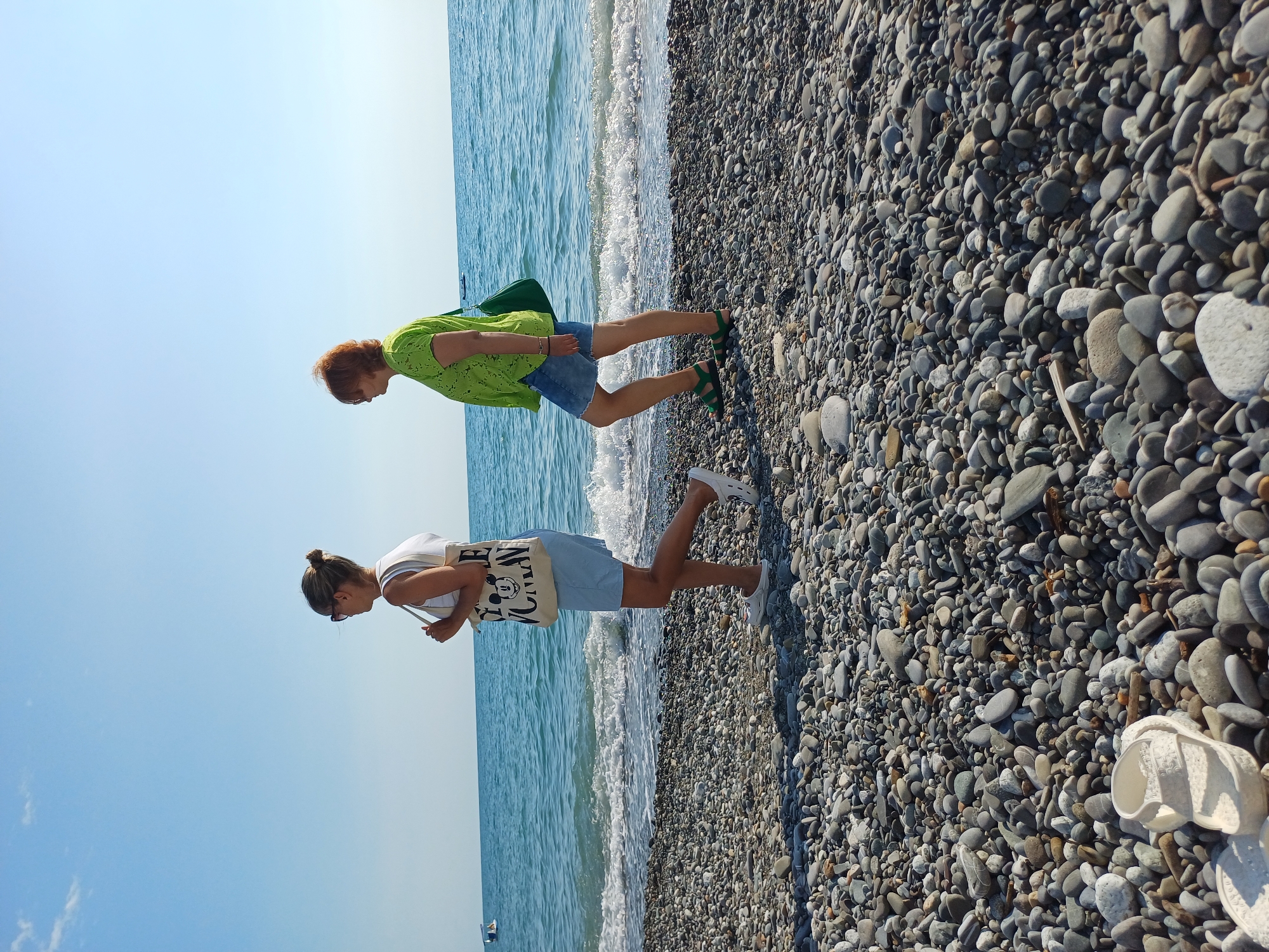 Two women are on a pebble beach, with the sea on their left and a clear blue sky on their right. The image is oriented vertically, so the women appear to be walking horizontally.
The woman in the foreground is wearing a white tank top, light blue shorts, and white slip-on shoes. She has her hair tied up in a bun and is wearing sunglasses. She is carrying a beige tote bag with a print of Mickey Mouse and the words "Oh Mickey, you're so fine!" She is looking down towards the beach.
The woman in the background is wearing a bright lime green patterned top and denim shorts, with green sandals. She has short, reddish-brown hair and is carrying a green bag over her shoulder. She is also looking downwards.
The beach is covered in small, smooth grey pebbles. Gentle waves are lapping at the shore. The lighting suggests it is daytime, likely sunny. There is a white sandal visible on the pebbles in the bottom right corner of the image.