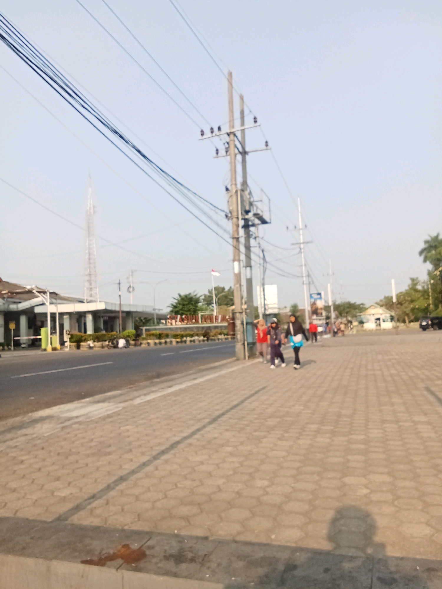 This image depicts a street scene in Kaliwungu, Indonesia, likely during daytime given the lighting. The foreground features a light-colored, hexagonal-patterned paved sidewalk adjacent to a road. A concrete barrier runs along the edge of the sidewalk, with some debris visible on it.
In the mid-ground, several individuals are walking on the sidewalk. Three women are walking together, with one wearing a black headscarf and a light blue bag, another in a dark purple outfit and headscarf, and a third in a red top and headscarf. Further down the sidewalk, other people are visible, some appearing to be walking or standing.
To the left of the road is a building complex with a white facade and a covered walkway. A tall, lattice-like tower, possibly a cell tower, stands behind this building. A flag is visible on a flagpole near a sign that appears to read "STASIUN K...". The area is lined with trees and shrubs.
In the background, more buildings and vegetation are present, including palm trees. Power lines and utility poles are prominent features of the scene, stretching across the sky. The sky is a pale blue, indicating clear weather. A black car is parked on the right side of the road in the distance. The overall impression is of a functional urban or semi-urban environment.