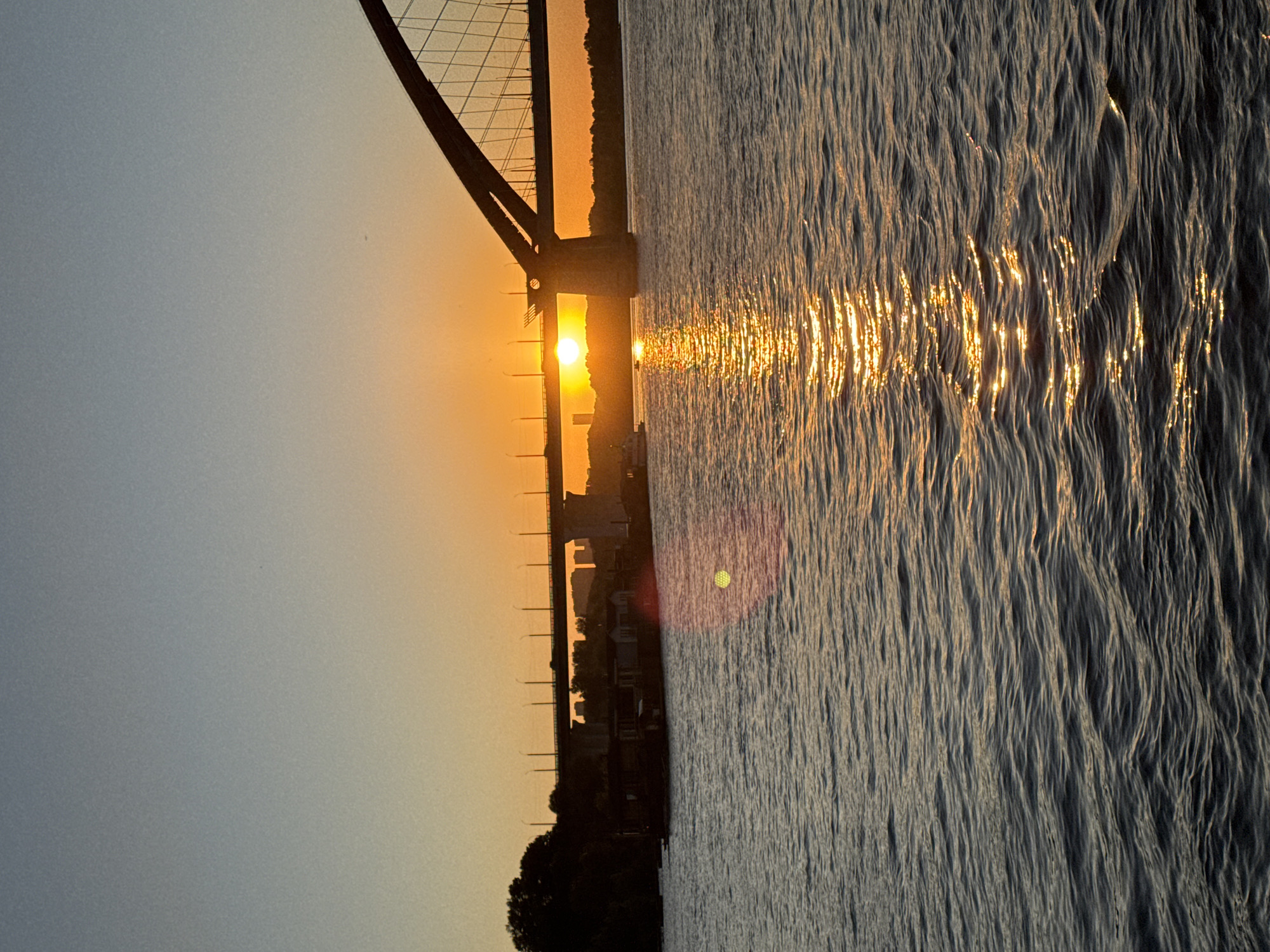 This is a vertical, medium-distance photograph of the sunset over the Ob River in Novosibirsk, Russia. The sun is setting behind a large arched bridge and the water reflects the golden light. The sky is a gradient of orange and blue. A few boats are visible on the water, as well as buildings and trees on the far bank. The image captures a serene and picturesque scene of an urban sunset.