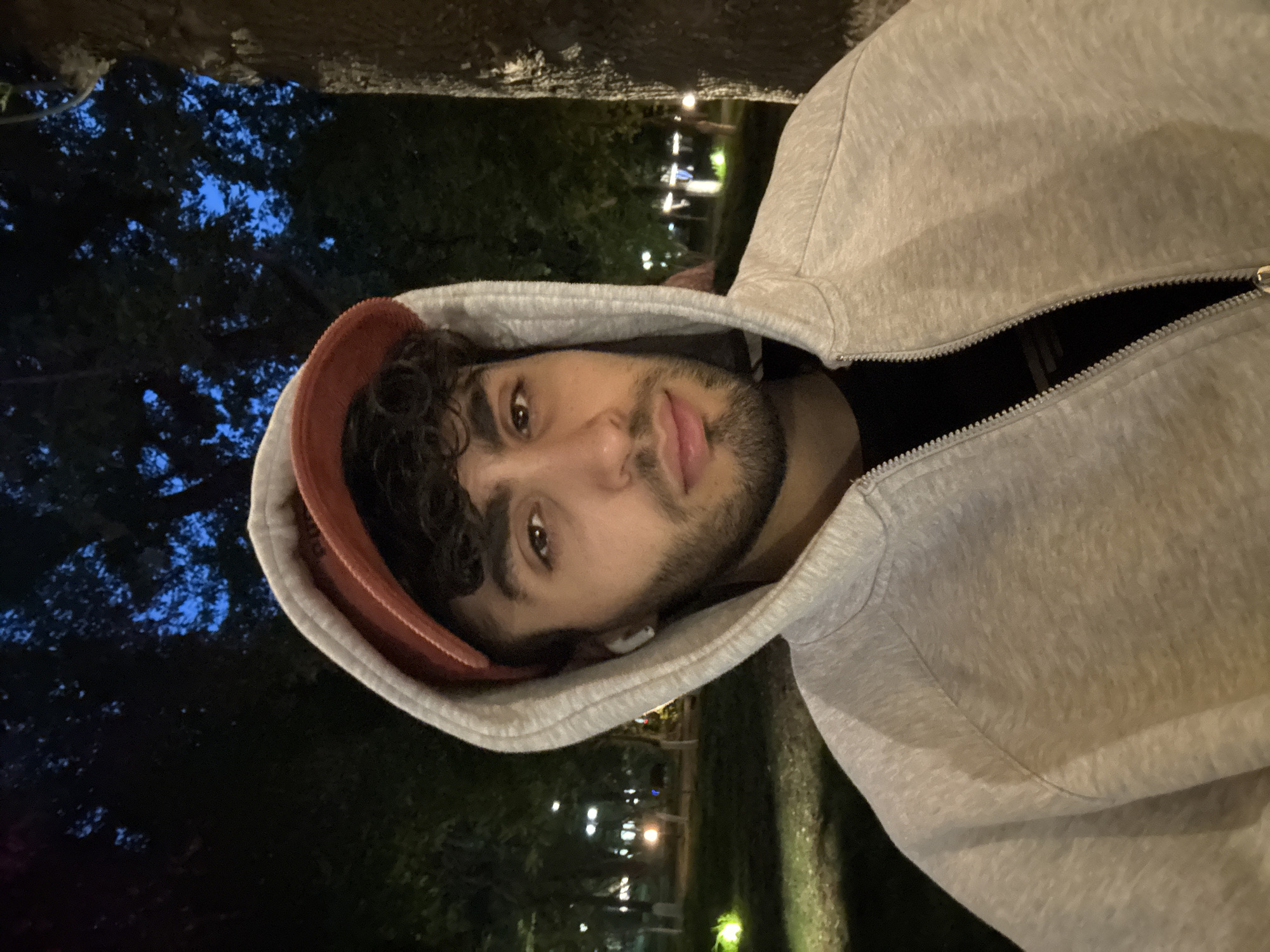 A young man with brown hair and a beard is wearing a grey hoodie and a red hat. He is in Yerevan, Armenia, in a park at night, with trees and streetlights visible in the background.