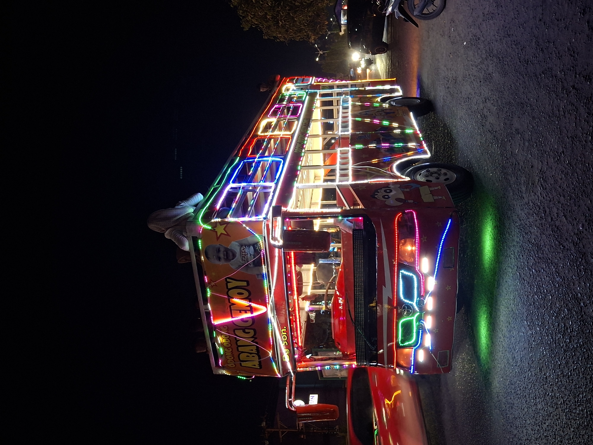 A vibrant, brightly lit modified vehicle, possibly a bus or truck, is parked on a dark asphalt surface at night, likely a street, within the city of Lhokseumawe, Indonesia. The vehicle is heavily adorned with colorful LED lights outlining its structure, and various decorative elements including cartoon characters, and an image of a baby on a billboard with text "ABANG GEMOY" beside it. In the background, there are glimpses of other vehicles, streetlights, and foliage suggesting an urban setting. The wet pavement is reflective, amplifying the effect of the colorful lighting.