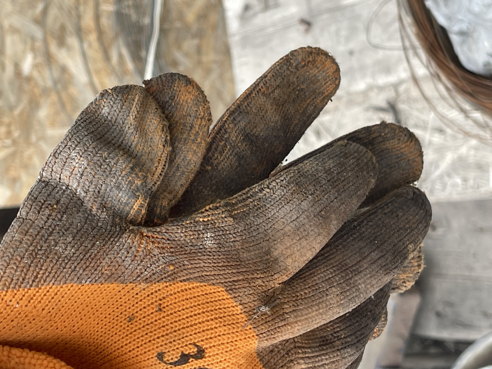 A close-up shot shows a gloved hand, covered in dirt and grime, resting on a surface. The glove is primarily orange and brown, with a knitted texture that appears worn and stained. Some of the dirt has a reddish-brown hue, suggesting it's from soil or rust. The fingers of the glove are curved, indicating the hand is in a relaxed or loosely gripping position. The background is blurred, but appears to be an outdoor environment with a wooden texture and some metallic elements, possibly a construction site or workshop. The image was taken in Bishkek, Kyrgyzstan.