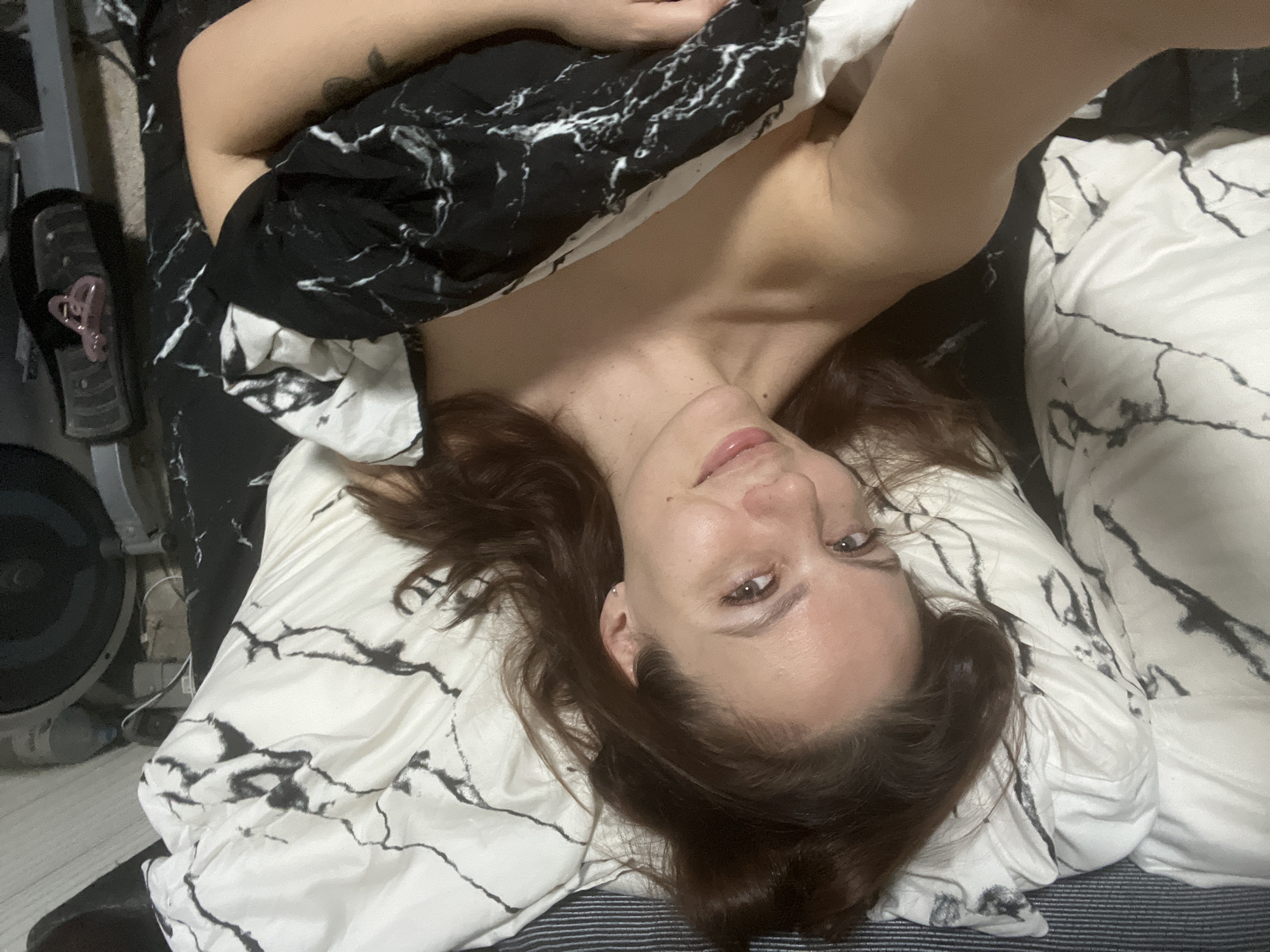 In a self-portrait taken from an overhead perspective, a young woman lies on her back, looking directly into the camera with a relaxed expression. Her head is slightly tilted, and her lips are parted in a subtle smile. She has dark, wavy hair spread around her head on the pillow. Her fair skin is visible on her shoulders and upper chest, suggesting she is covered by bedding. A small piercing is visible on her left ear, and a faint tattoo is seen on her upper right arm.
She is lying on a white pillow adorned with black, marble-like patterns, matching the duvet or blanket that partially covers her body. The bedding creates a stark contrast between black and white, adding a graphic element to the scene. To the left of the bed, a dark object resembling part of an exercise machine, possibly a stationary bike or elliptical, is visible. A pink hair clip or accessory rests on it. Wires are also visible near this equipment, suggesting an indoor setting like a bedroom. The floor or surface beneath the equipment appears to be light-colored or striped. The lighting is ambient, without strong indications of natural daylight. The overall mood is intimate and casual, capturing a moment of repose. This scene is set in Chaikovsky, Russia. There is no visible text in the image.