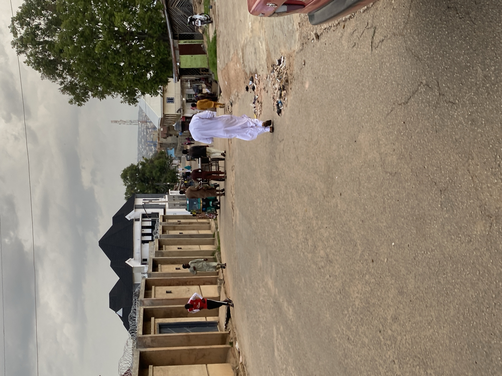 The image depicts a street scene in Jos, Nigeria, under an overcast sky. In the foreground, a paved road shows some wear and tear, with scattered debris along the right edge. A red car is partially visible on the far right. Several individuals are walking on the road and sidewalk.
On the left side of the road, a modern building with a dark, multi-peaked roof is under construction, bordered by a tan-colored wall topped with barbed wire. A person in a red and white athletic outfit walks away from the camera towards this building. Further down the street, another person in light-colored traditional attire walks in the same direction.
The mid-ground features more buildings of varying styles, some appearing older and more utilitarian. A green auto-rickshaw is parked on the road, with people gathered around it. A few market stalls or small shops with corrugated metal roofs are visible on the right side.
In the background, a transmission tower rises above the cityscape, and the distant horizon is marked by more buildings and a hazy sky. A large, leafy green tree stands prominently on the right side of the image, partially obscuring the background. The overall impression is of a busy, developing urban environment.