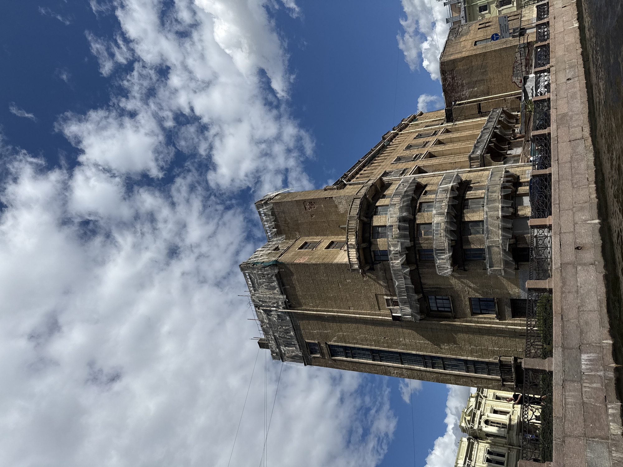 The image shows a historic brick building with a distinctive architectural style, situated along a canal in Saint Petersburg, Russia. The building is tall and rectangular, with multiple floors featuring numerous windows. Several balconies with intricate railings project from the facade. The upper portion of the building is covered with protective sheeting, suggesting ongoing renovation or maintenance work.
In the foreground, a granite embankment lines the canal, with a black wrought-iron fence along its edge. Greenery is visible behind the fence. The water in the canal is dark and appears calm.
The sky above is a vibrant blue, dotted with fluffy white clouds. To the left of the main building, other historical buildings with ornate facades and windows can be seen. A person is standing on the embankment, facing away from the camera, looking towards the canal. The overall scene conveys a sense of historical grandeur and urban life in Saint Petersburg.