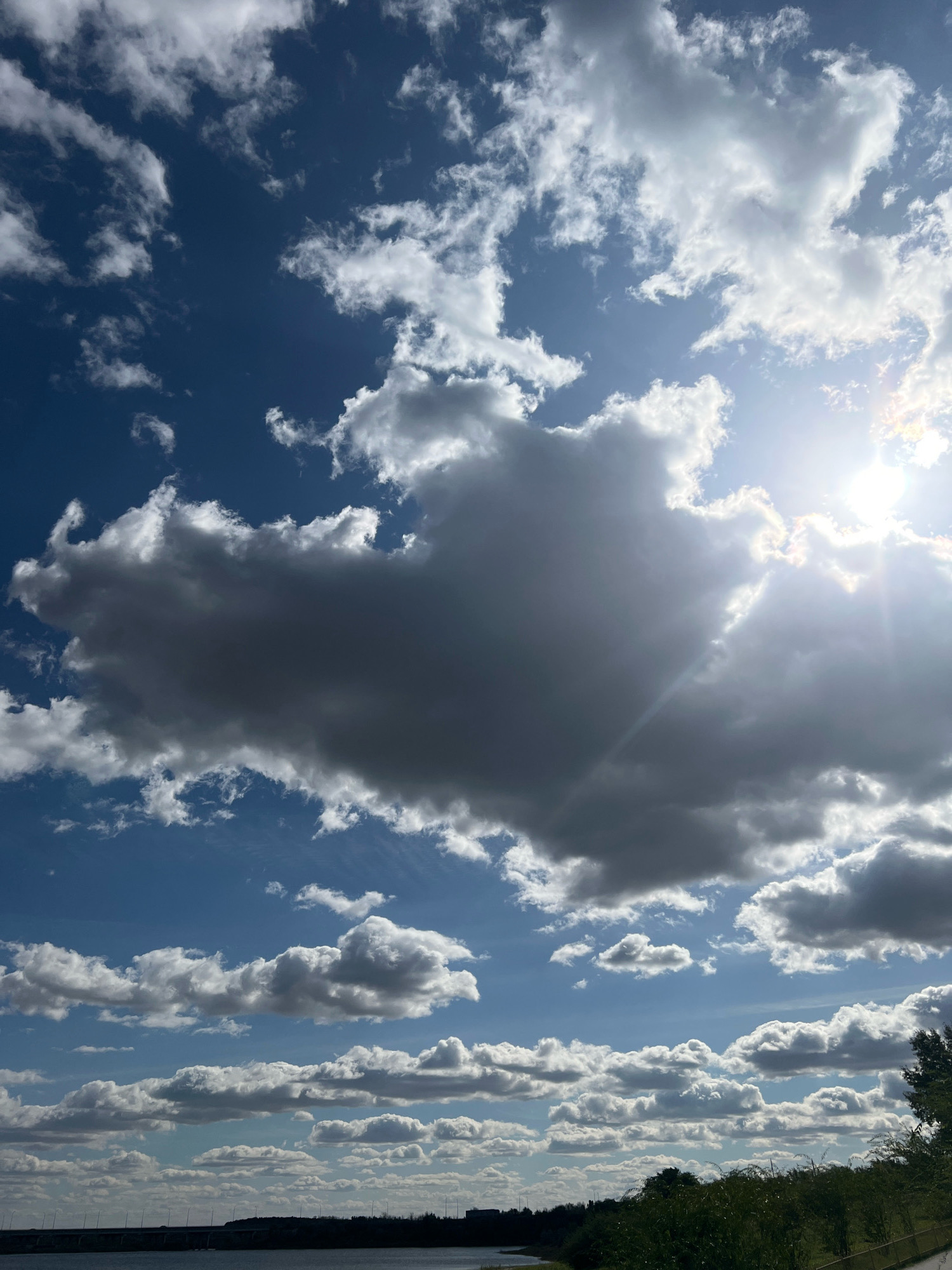 The image displays a bright, partly cloudy sky over a body of water. The sun is visible in the upper right quadrant, partially obscured by fluffy white clouds that transition to darker grey clouds in the center of the frame. The sky is a deep blue. In the lower portion of the image, the horizon shows a body of water with a distant bridge and silhouetted vegetation along the bank. The scene appears to be a natural landscape, possibly a reservoir or large lake. The location is identified as the Tobol River embankment in Kostanay, Kazakhstan. There are no people or visible actions taking place in the image.