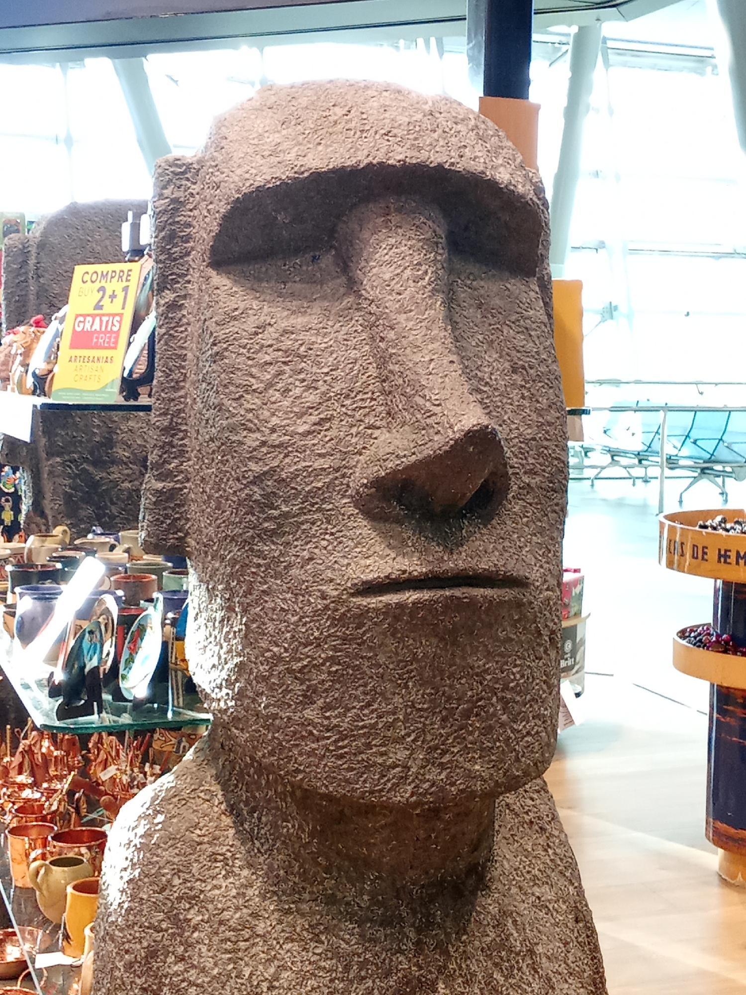 The image shows a close-up view of a large stone Moai statue, a replica of those found on Easter Island. The statue is positioned in the foreground, occupying most of the frame. Its rough, textured surface is clearly visible, with details of its face including the large nose, deep-set eyes, and a stern mouth.

Behind the statue, to the left, a sign is visible with the text "COMPRE BUY 2+1 GRATIS FREE ARTESANIAS ARTISAN CRAFTS". This suggests the statue is located in a shop or market selling crafts. Various other craft items, including pottery and figurines, are displayed on shelves and tables in the background, partially obscured by the Moai.

Further in the background, through a large glass window, a seating area with chairs and tables is visible, resembling an airport waiting lounge or a public space within a building. The overall environment appears to be indoors, with bright, diffused lighting.

The location is specified as the city of Pudahuel, Chile. Given the presence of what appears to be an airport lounge and souvenir shops selling local crafts, it is highly probable that this scene is within the Arturo Merino Benítez International Airport (SCL) in Pudahuel, which serves Santiago.