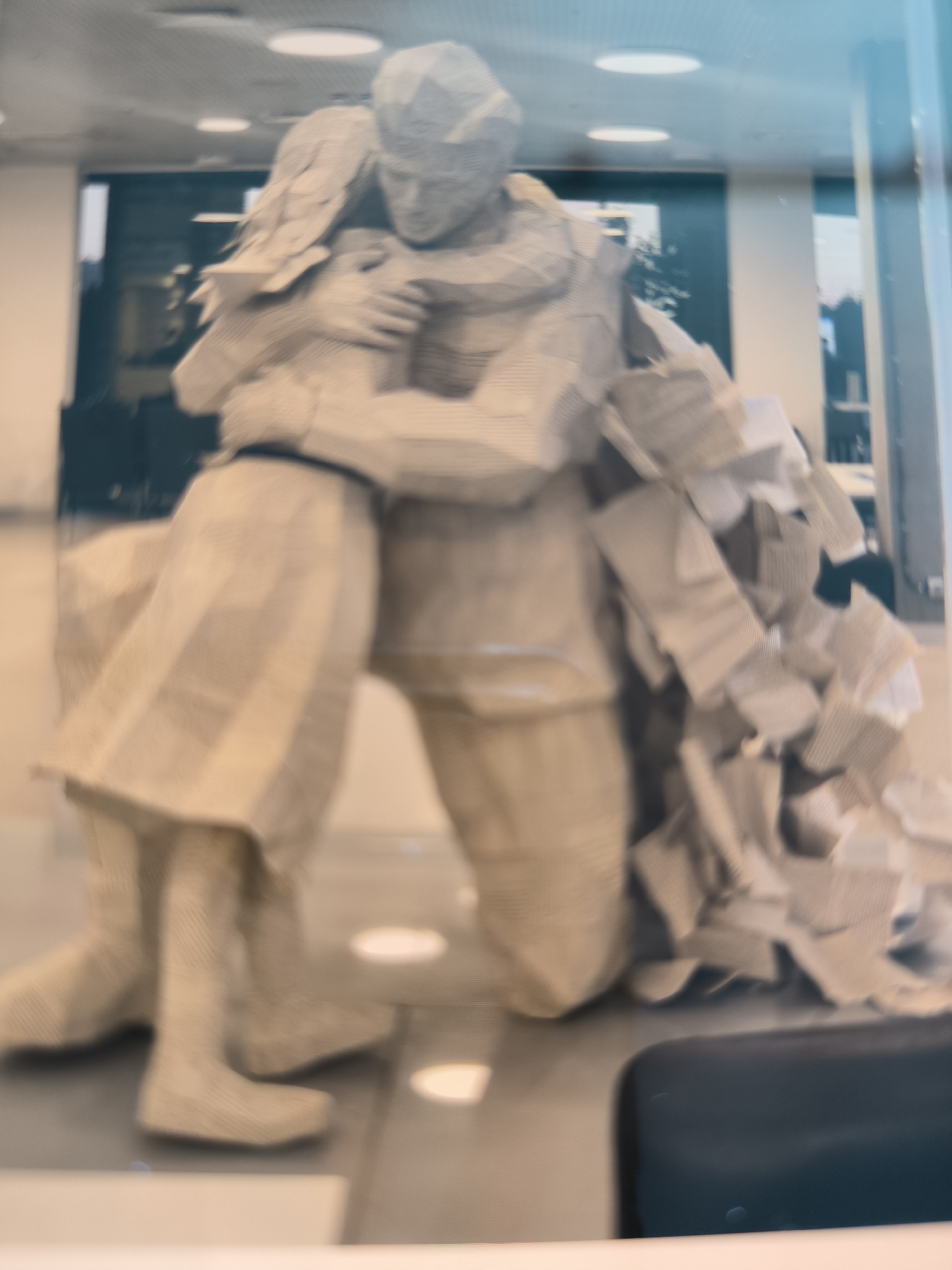 The image shows a blurred sculpture made of paper, depicting two figures embracing. The figures are standing, and one appears to be kneeling while hugging the other. The sculpture is light-colored and appears to be made from folded and rolled paper, with some areas resembling newsprint. The background is out of focus but suggests an indoor setting with lighting fixtures on the ceiling and glimpses of what might be office furniture or displays. The overall impression is of an artistic installation. The location in the city of Oktyabrsky, Russia, is not evident from the image itself.