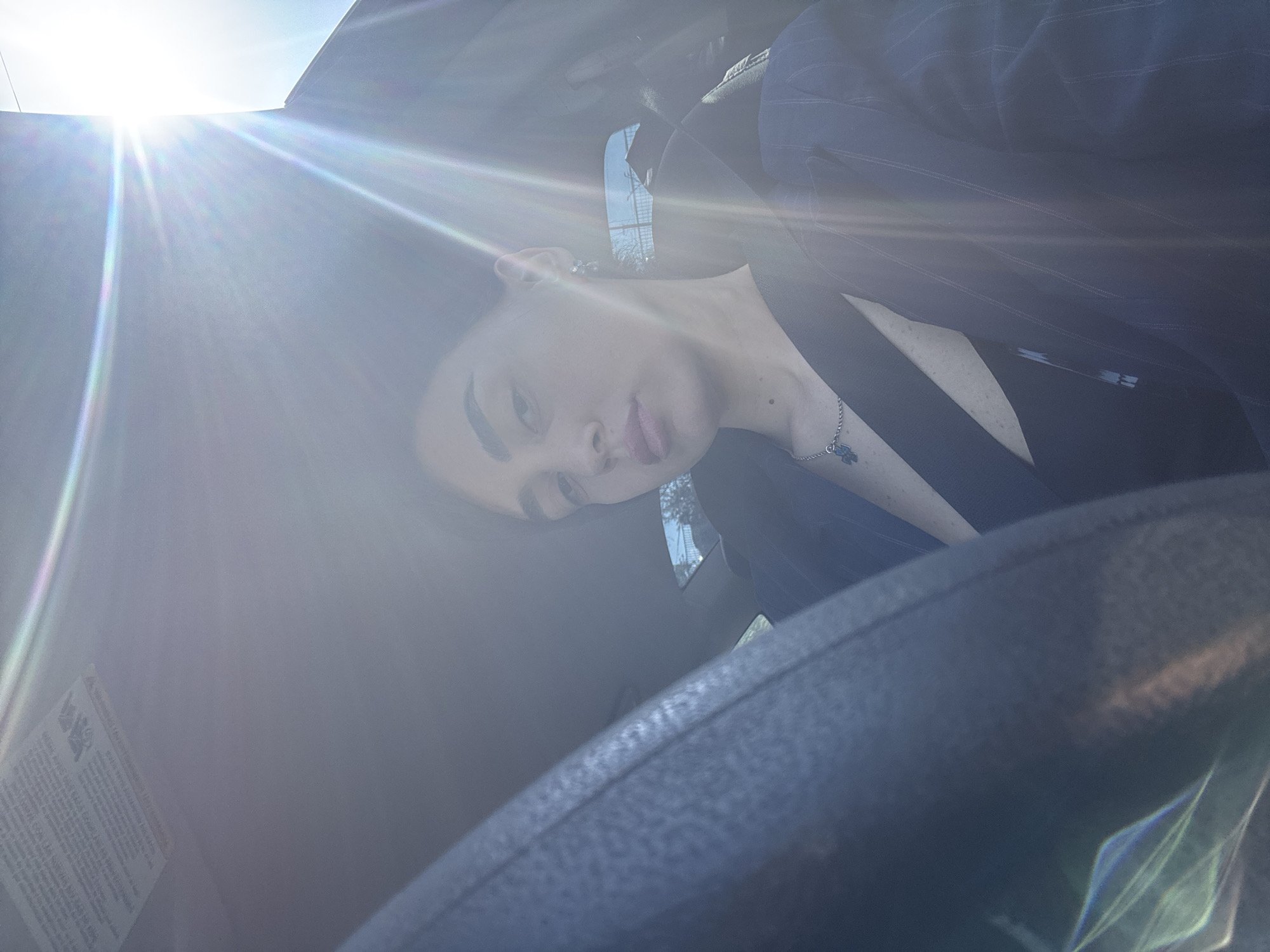 A close-up photograph taken from inside a vehicle, likely a car, shows a person with pale skin, dark eyebrows, and dark hair. The person wears a dark pinstripe blazer over a light-colored shirt and a silver necklace with a blue charm. The person's head is tilted towards the camera. The interior of the car is predominantly dark, with the steering wheel visible in the foreground. A bright sunlight flare is present in the upper left corner of the frame. A sticker with text is visible near the lower left corner, likely a manufacturer's warning label. Through the car windows, glimpses of a street and building can be seen, but are too blurred to determine their location. The city of Tbilisi, Georgia cannot be verified from this image alone.