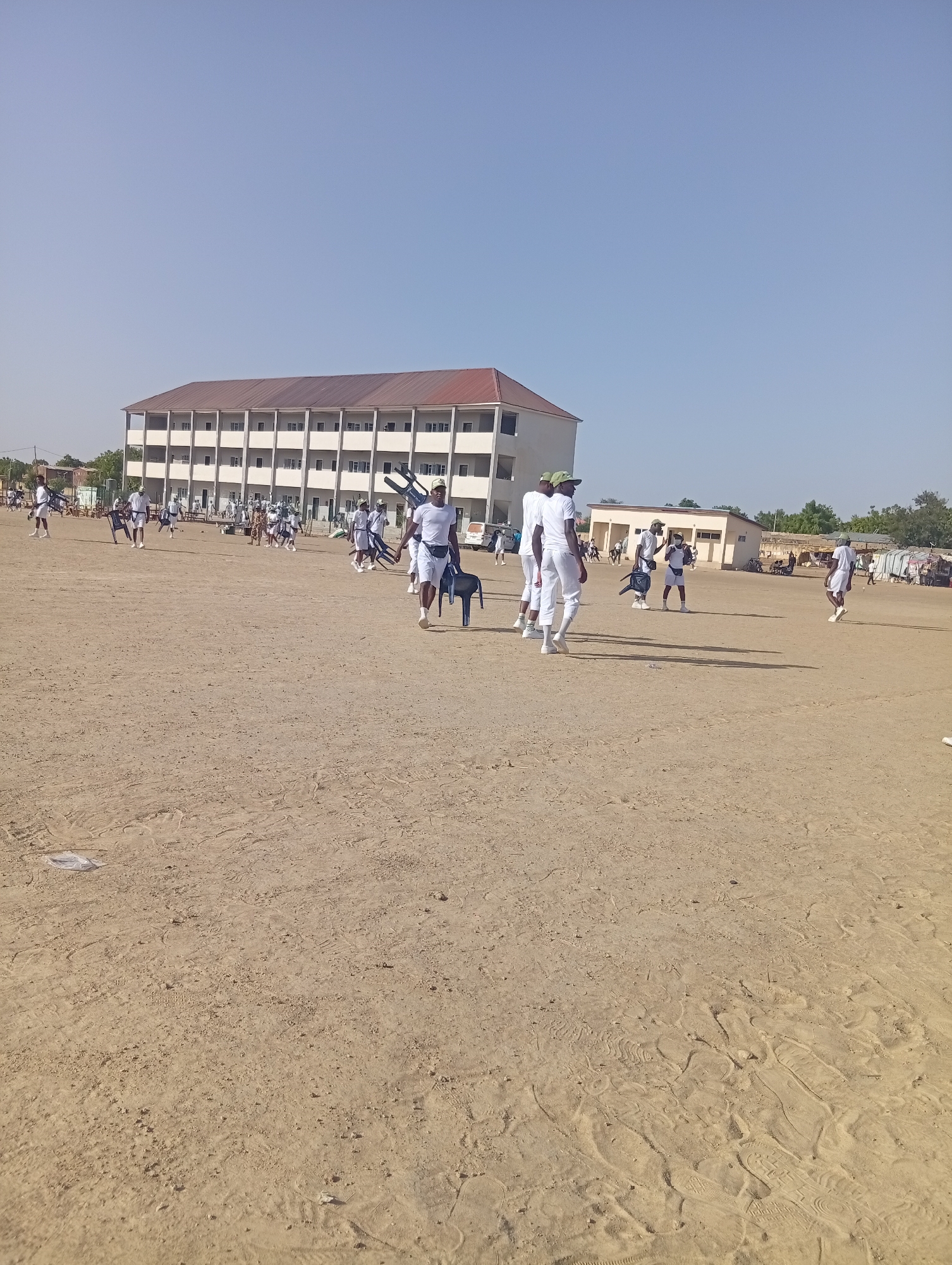 The image shows a large, flat, dirt field with numerous people scattered across it. In the background is a multi-story building with a red roof and white walls, suggesting an institutional structure. Most of the people are wearing white uniforms and some are carrying chairs. The sky is clear and light blue, indicating a sunny day. There is sparse vegetation along the edges of the field, and some smaller buildings can be seen in the distance. The location is likely Maiduguri, Nigeria. People are interacting by walking across the field, some of them carrying chairs.
