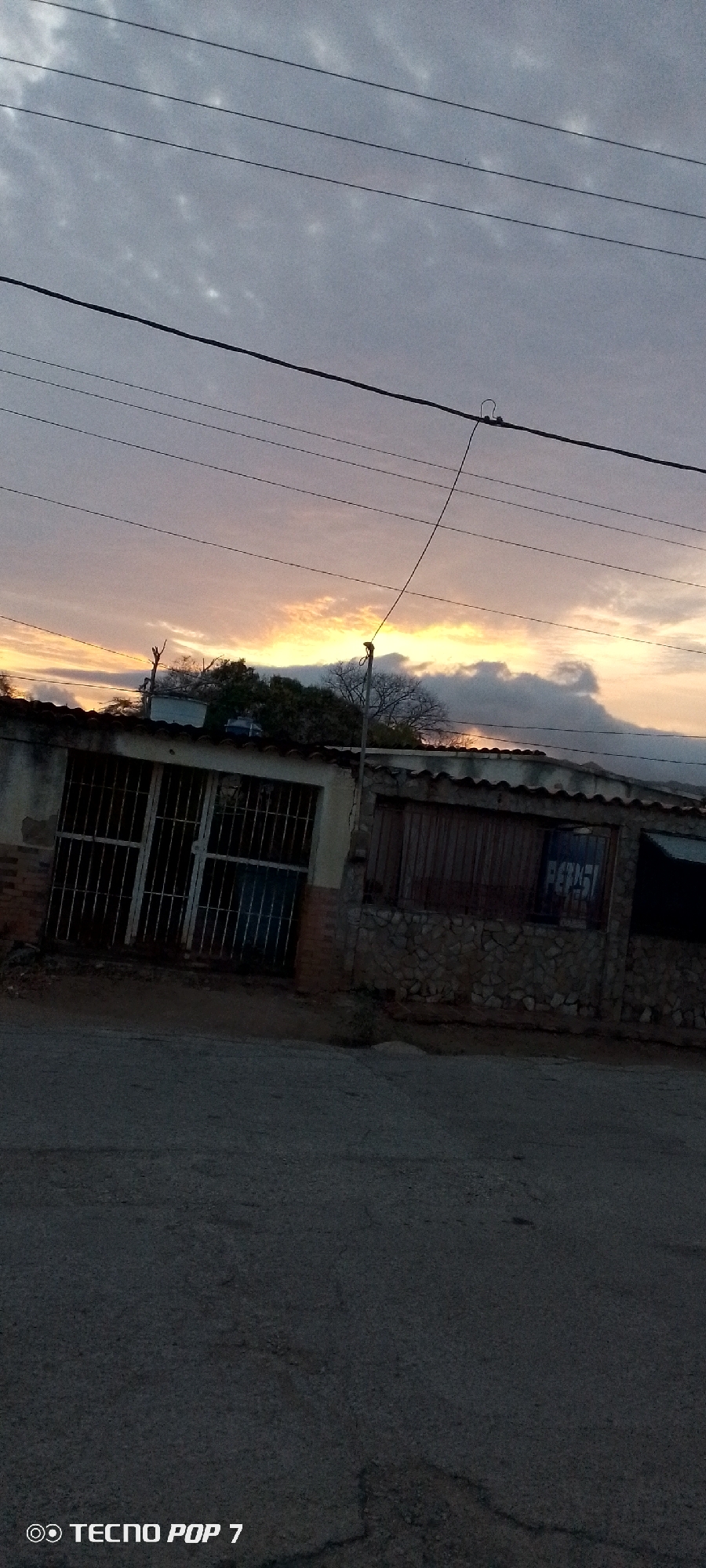 The image captures a street scene in what is likely Altagracia, Venezuela, based on the metadata. The sky is filled with clouds tinged with the orange hues of either sunset or sunrise. Several power lines stretch across the upper part of the frame.
In the middle ground, there is a single-story building. One section features a metal-grated gate, while the adjacent section has wooden boards and stonework. A faded Pepsi sign is visible on a section of the building.
The foreground is dominated by the paved road, which appears dark, with visible cracks and imperfections. The camera's model, a "Tecno Pop 7," is watermarked in the bottom left corner. There are no people visible in the shot. The environment suggests a residential or commercial area in a state of slight disrepair.