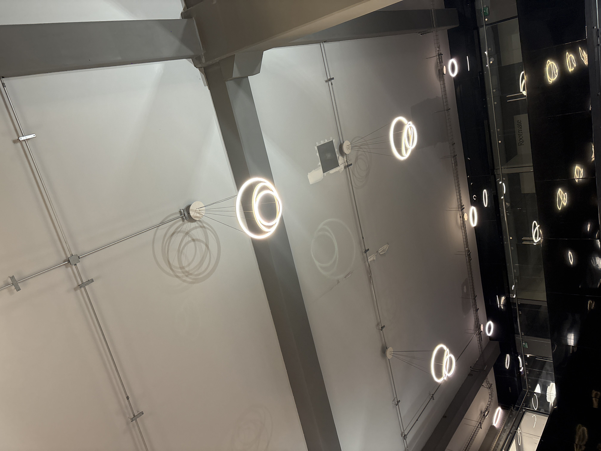 The image presents an upward view of a modern interior space, likely a commercial or public building in Puebla, Mexico. The ceiling is primarily white with visible structural beams and conduits running across. Three illuminated circular light fixtures are prominently displayed, suspended from the ceiling by wires. The light fixtures cast faint shadows on the ceiling.
To the right of the frame, a dark, reflective surface is visible, showing mirrored images of similar circular light fixtures and the word "Rootmale". This surface is likely a black glass wall or window, suggesting the location is within a building.
The lighting is bright, and the scene appears unoccupied by people. The overall impression is of a clean, modern architectural space.