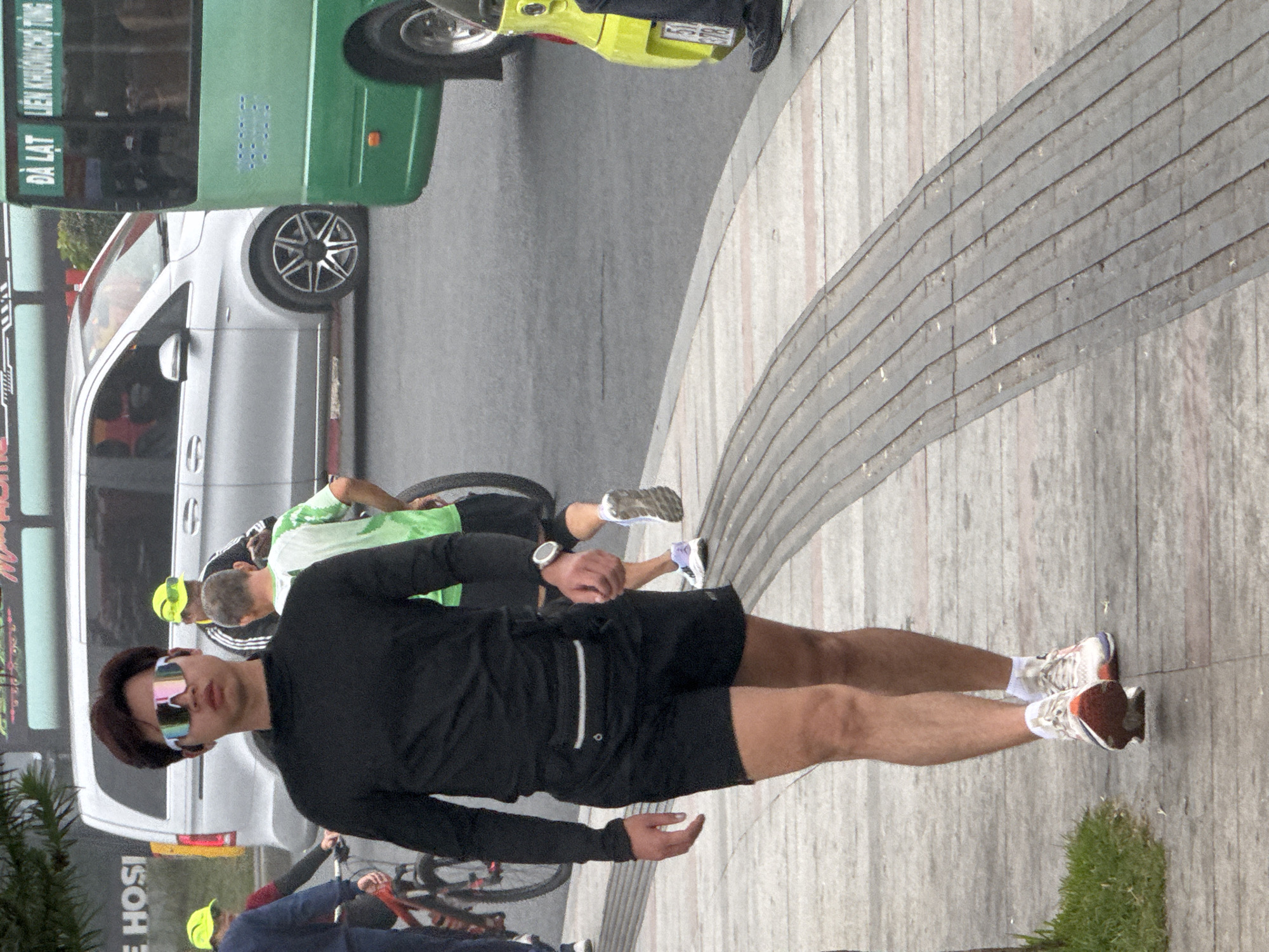 A young man wearing sunglasses, a black long-sleeved shirt, and black shorts walks along a paved sidewalk in Da Lat, Vietnam. He has a fanny pack around his waist and is wearing athletic shoes. Behind him, a silver minivan and a green bus are visible on the street, along with other pedestrians and cyclists. The weather appears to be overcast. The man is walking away from the camera and his face is partially obscured by his sunglasses. The sidewalk has a patterned design of gray and pink tiles.