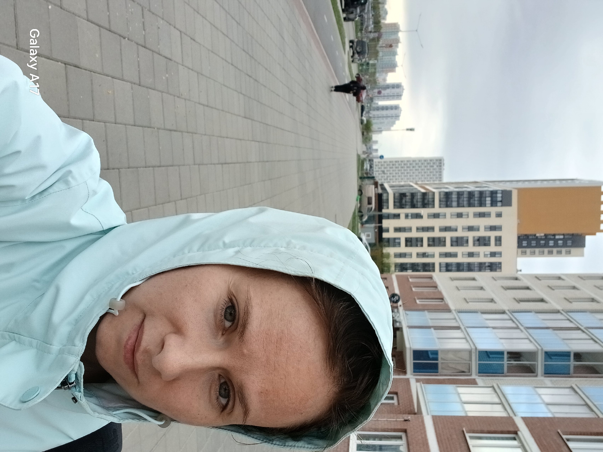 A selfie photograph shows a woman wearing a light blue hooded jacket standing on a brick sidewalk in Yekaterinburg, Russia. In the background, there is a street with cars and a person walking, followed by high-rise apartment buildings against a cloudy sky.