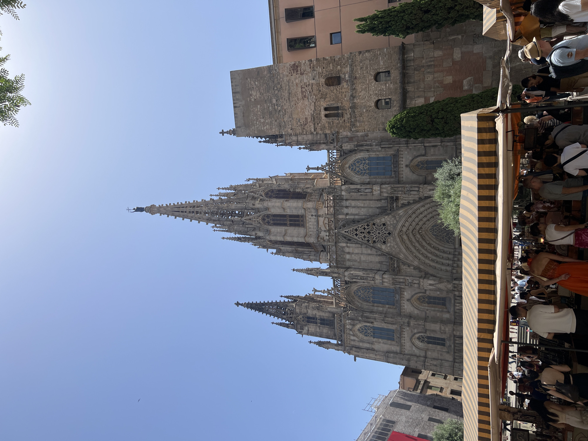The image shows the Barcelona Cathedral in Barcelona, Spain. It is a large Gothic cathedral with intricate stonework and tall spires. In front of the cathedral, there is a bustling outdoor market with many stalls and people browsing. The market stalls are covered with striped awnings. The sky is clear and blue, indicating a sunny day. The overall scene depicts a vibrant urban environment with a mix of historic architecture and lively commerce.