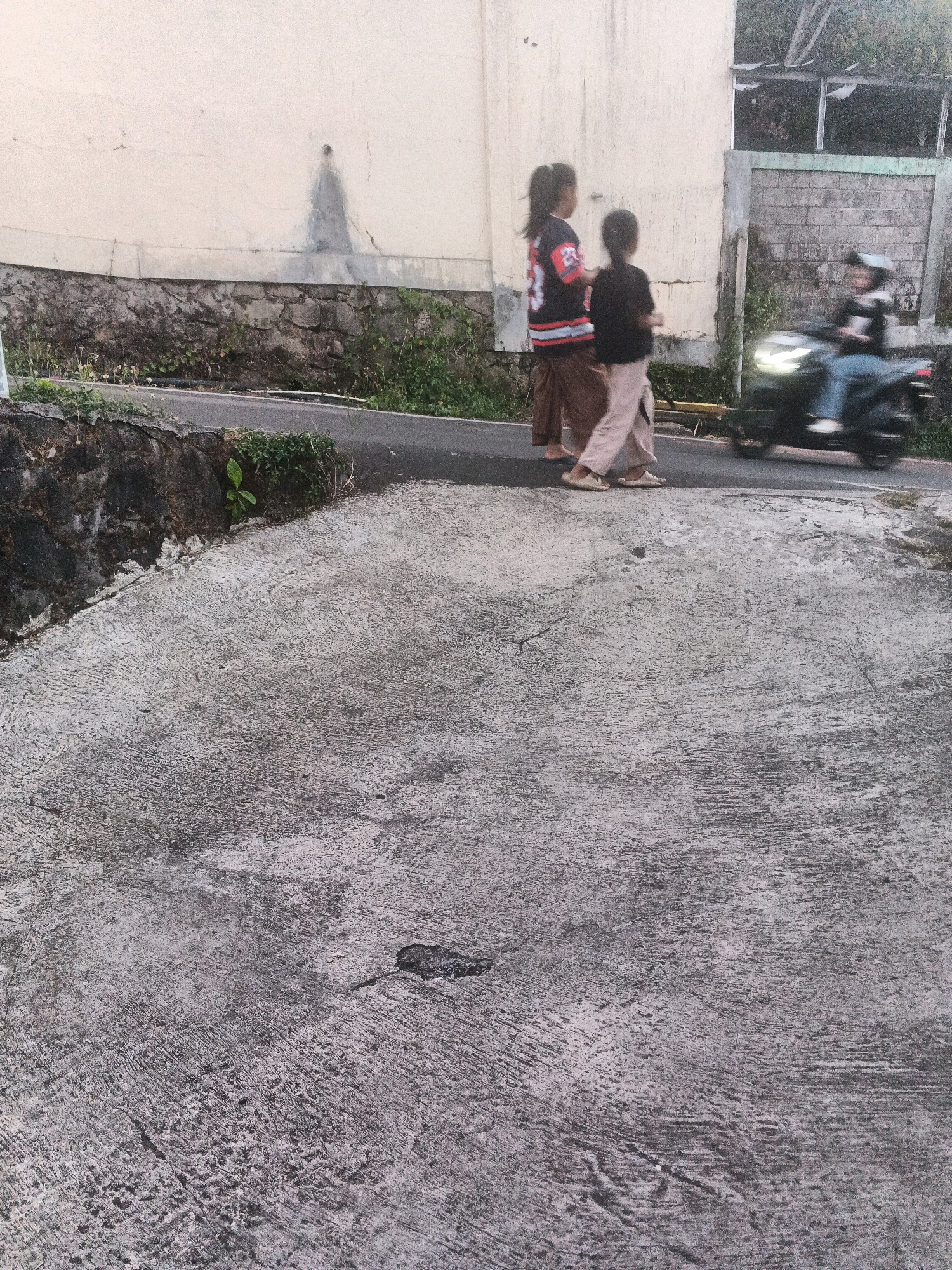 The image depicts a street scene in Keji, Indonesia. In the foreground, a textured concrete surface occupies the majority of the frame, appearing worn and uneven with visible lines and pits. A paved road runs horizontally across the mid-ground, bordered by a low, rough stone wall on the left and vegetation. Two young girls are walking on the road from left to right, one wearing a black and red jersey with the number 25 and the other a dark shirt. To the right of the girls, a person on a dark-colored motorcycle is riding on the road, moving away from the viewer and towards the right side of the image, with their head and upper body visible. The background features a light-colored wall, possibly of a building, with some faint dark markings. Further back, a brick wall and some greenery are visible. The overall lighting suggests it is daytime, possibly overcast.