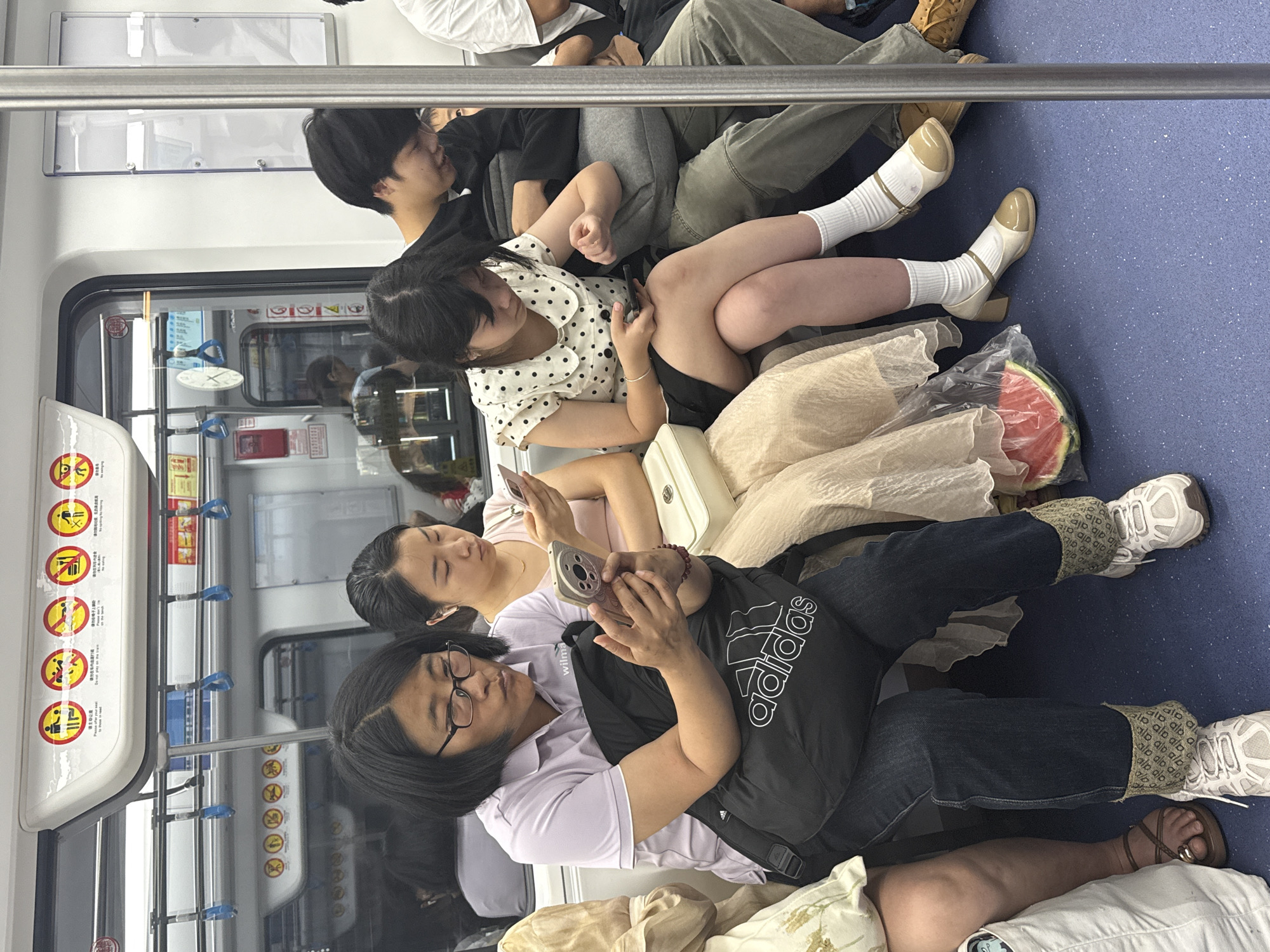 The image shows the interior of a subway car, with multiple individuals occupying the space. A woman with glasses and a white shirt is looking at her cell phone, which she is holding in her hands. Next to her, another woman is reclining on a pillow while using her cell phone. The subway car features warning signs and numerous handrails. In the front of the subway car are a group of younger people who are sitting together. Near one of them there appears to be half of a watermelon.